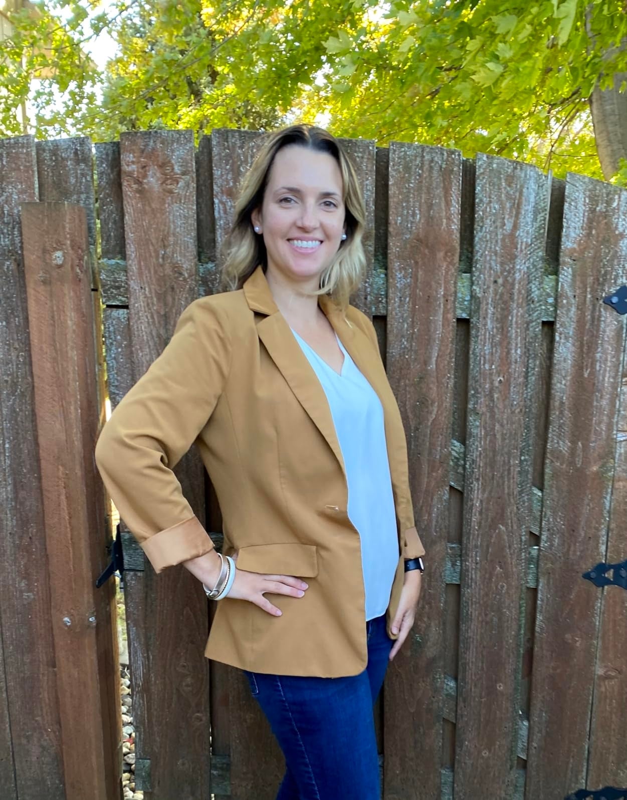 Person posing outdoors by a woody  fence, wearing a blazer, airy  blouse, and jeans, smiling