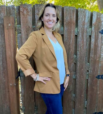 Person posing outdoors by a woody fence, wearing a blazer, airy blouse, and jeans, smiling