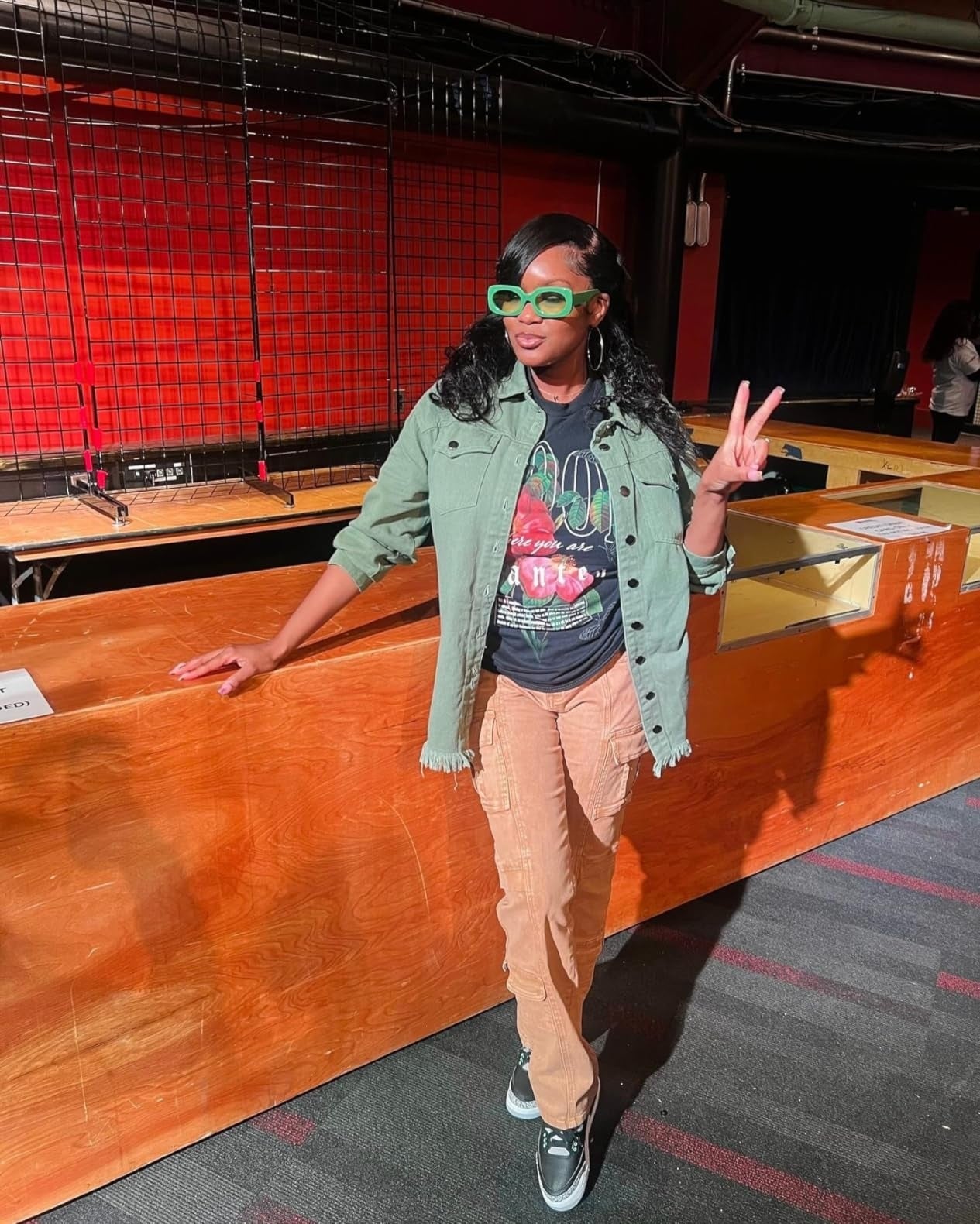 Person posing indoors wearing greenish  jacket, graphic tee, tan pants, and greenish  sunglasses, flashing a bid    sign