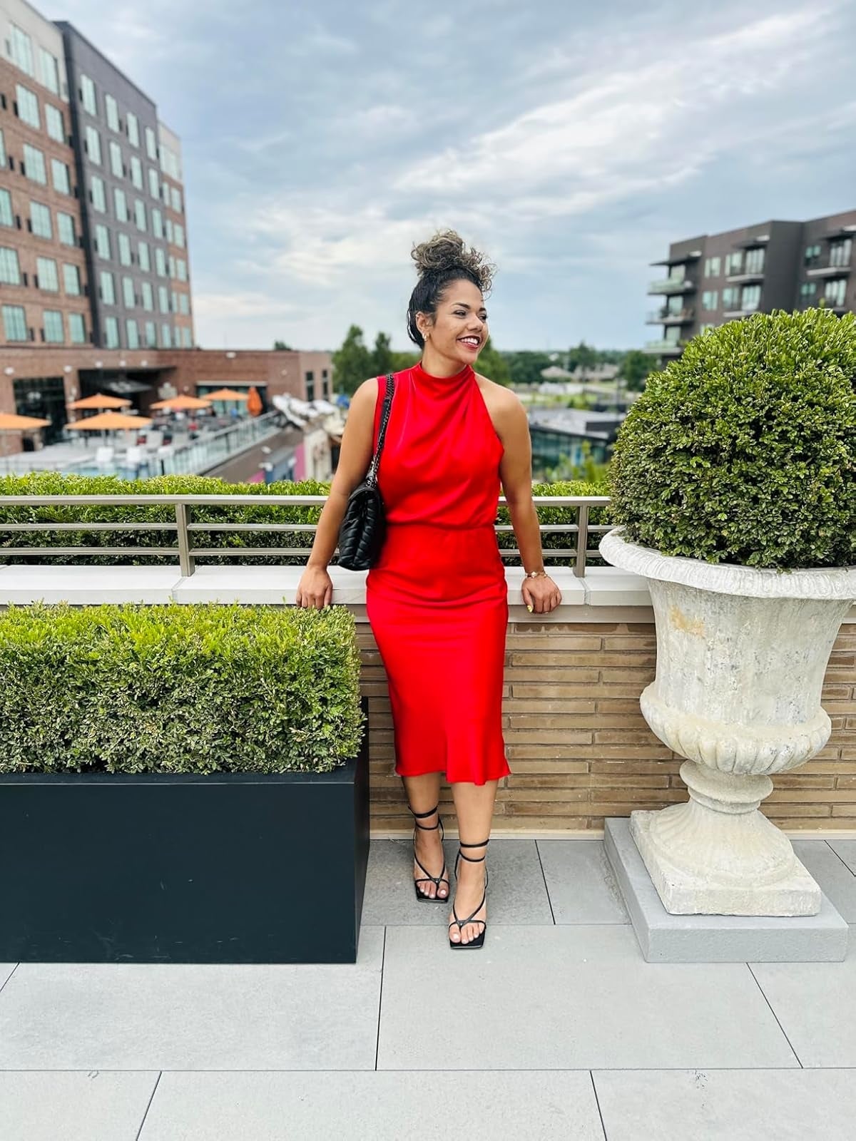 Person successful  a stylish sleeveless reddish  formal  with a halter neckline, lasting  connected  a rooftop patio, smiling and carrying a quilted handbag