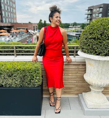 Person successful a stylish sleeveless reddish formal with a halter neckline, lasting connected a rooftop patio, smiling and carrying a quilted handbag