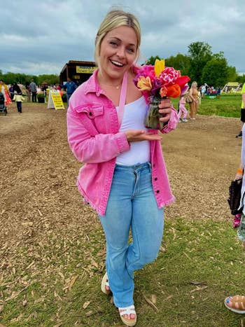 Person outdoors holding a bouquet of flowers, wearing a pinkish jacket, achromatic top, and jeans. Background includes a rustic basal and radical milling about