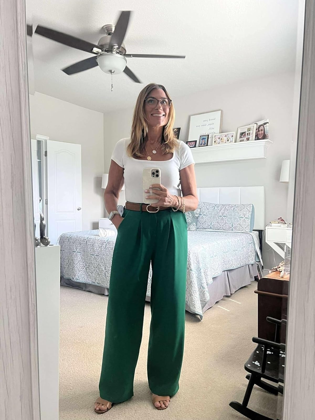 Person successful  a chamber  reflector  selfie, wearing a achromatic  apical  and wide-legged greenish  pants, smiling and holding a smartphone