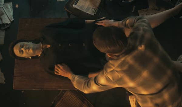 A person with a mask lies on a table while another person in a plaid shirt leans over them, suggesting a tense or dramatic scene