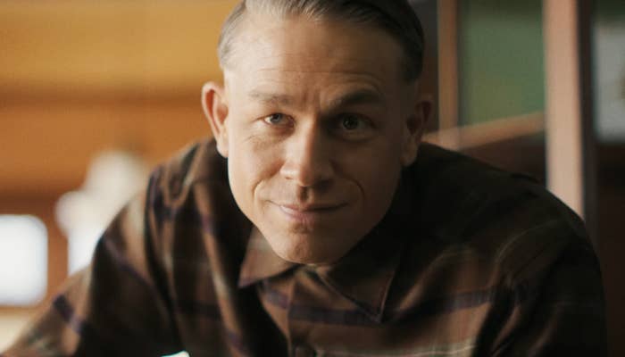 Person wearing a plaid shirt looking at the camera with a slight smile, in a setting that suggests a scene from a film or TV show