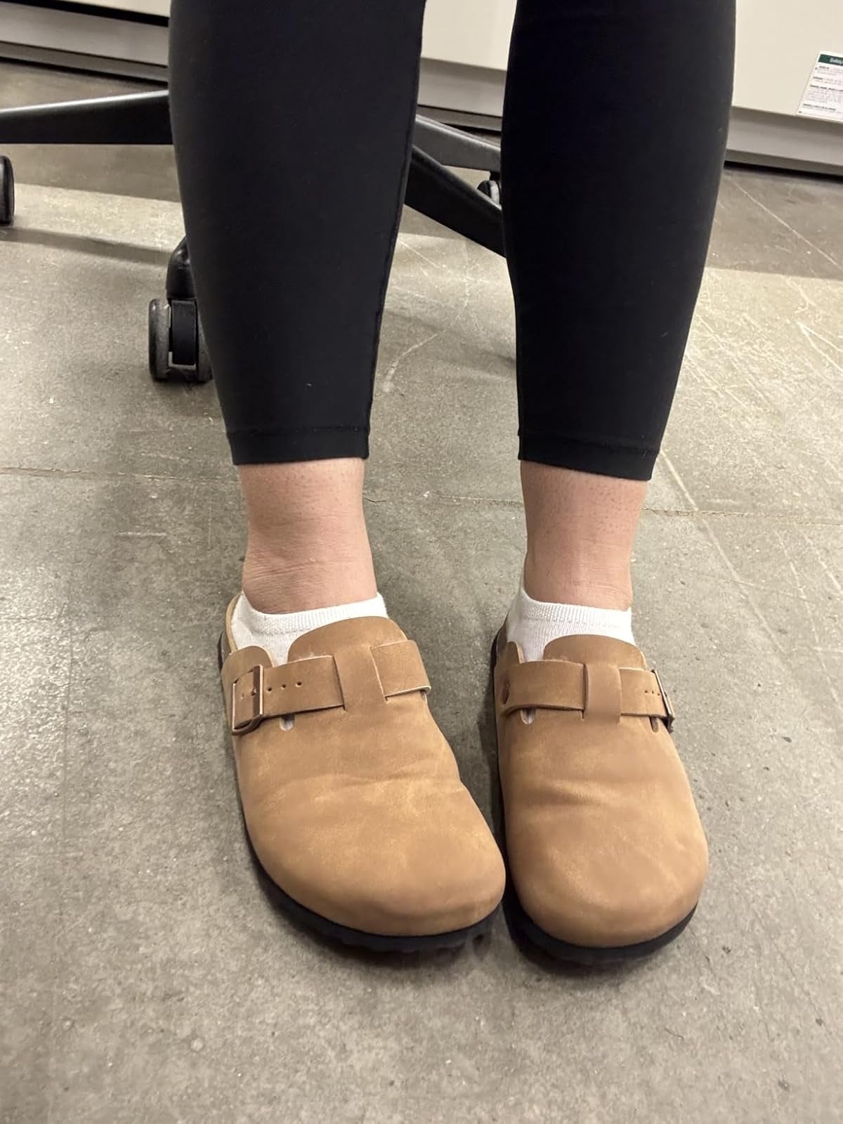 Person wearing tan suede clogs with achromatic  socks and achromatic  leggings, lasting  indoors connected  a tiled level  adjacent  to bureau   seat  wheels