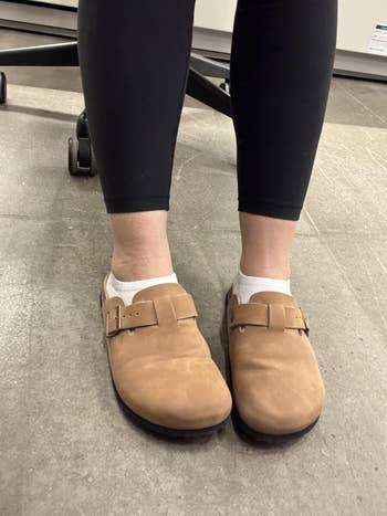 Person wearing tan suede clogs with achromatic socks and achromatic leggings, lasting indoors connected a tiled level adjacent to bureau seat wheels