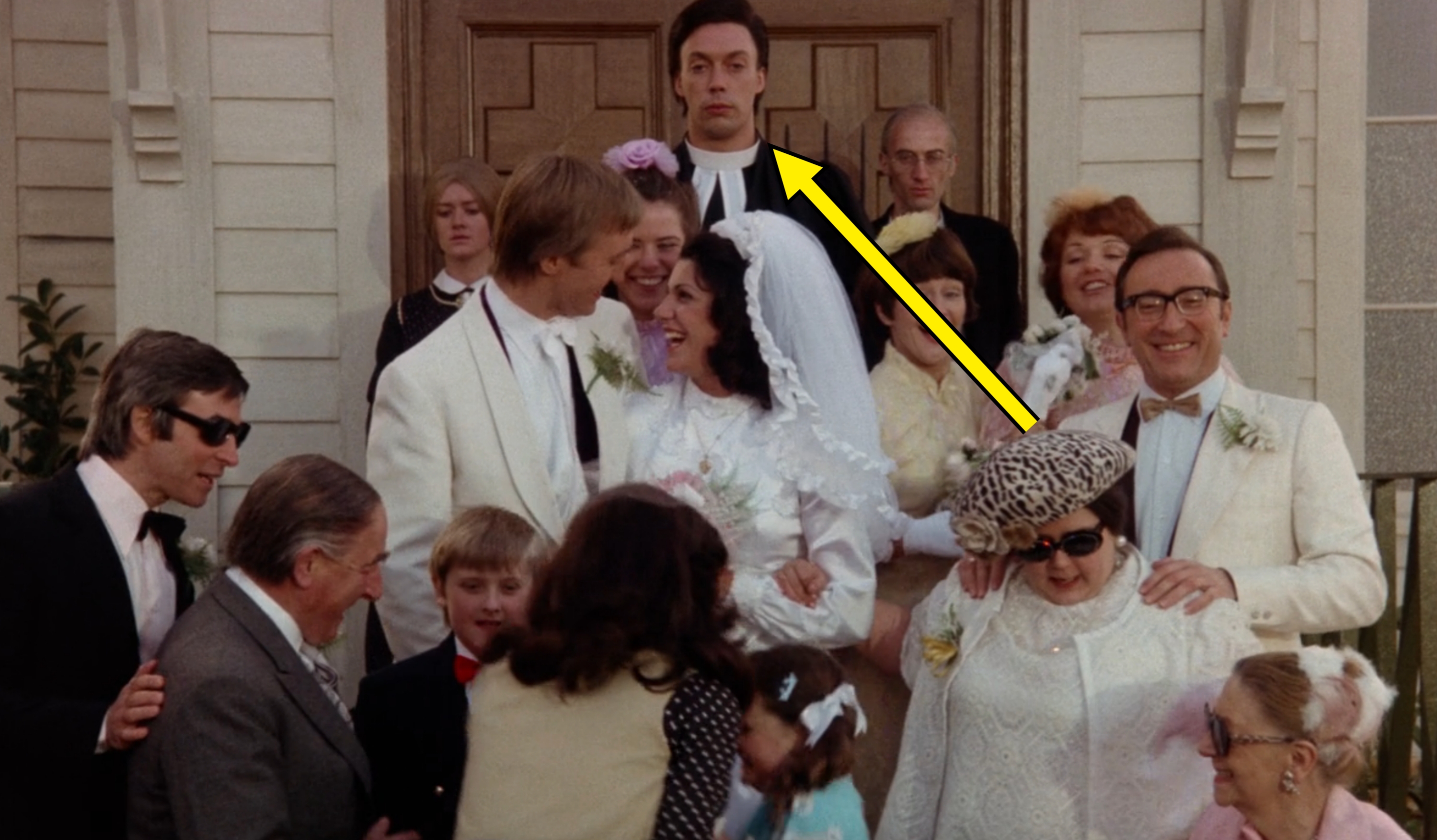Group of people at a wedding with a bride and groom smiling in the center, surrounded by guests in various outfits, including formal hats and suits