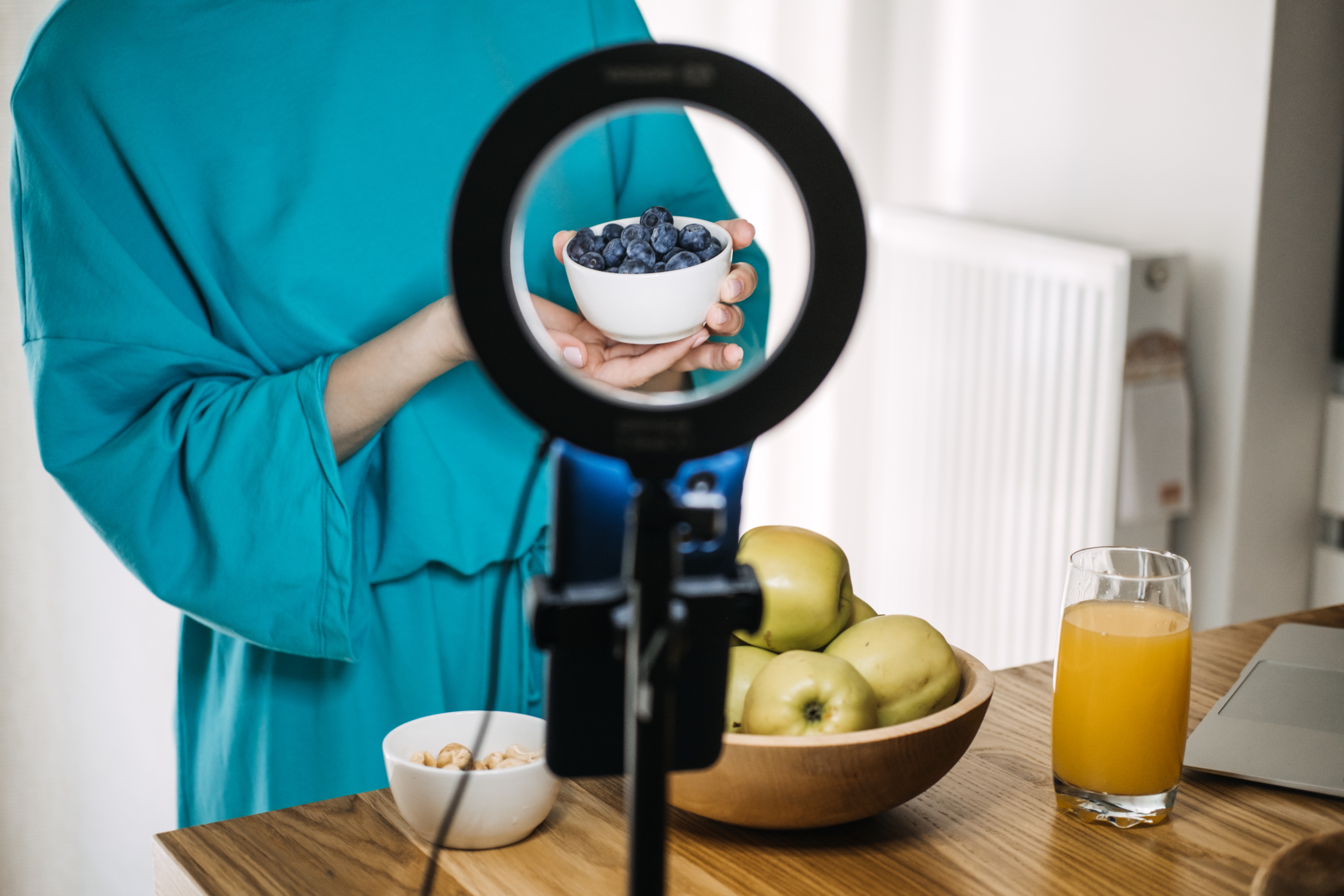 En person i et tealantrekk holder en skål med blåbær bak et ringlys. På bordet: Epler i en bolle, et glass juice og noen nøtter