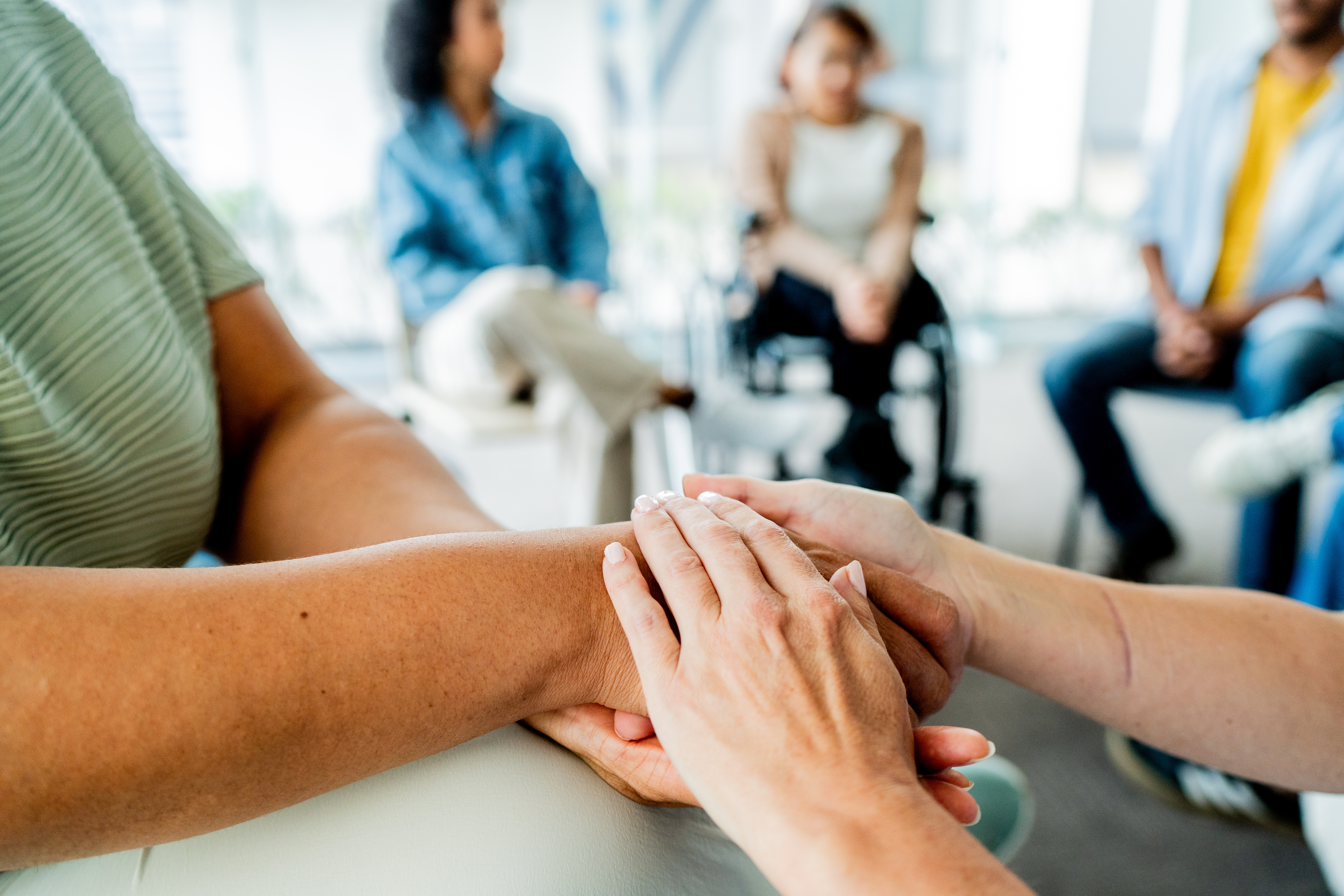 Folk som sitter i en sirkel, ett par holder hender i en støttende gest, noe som antyder et gruppemøte eller terapitimer
