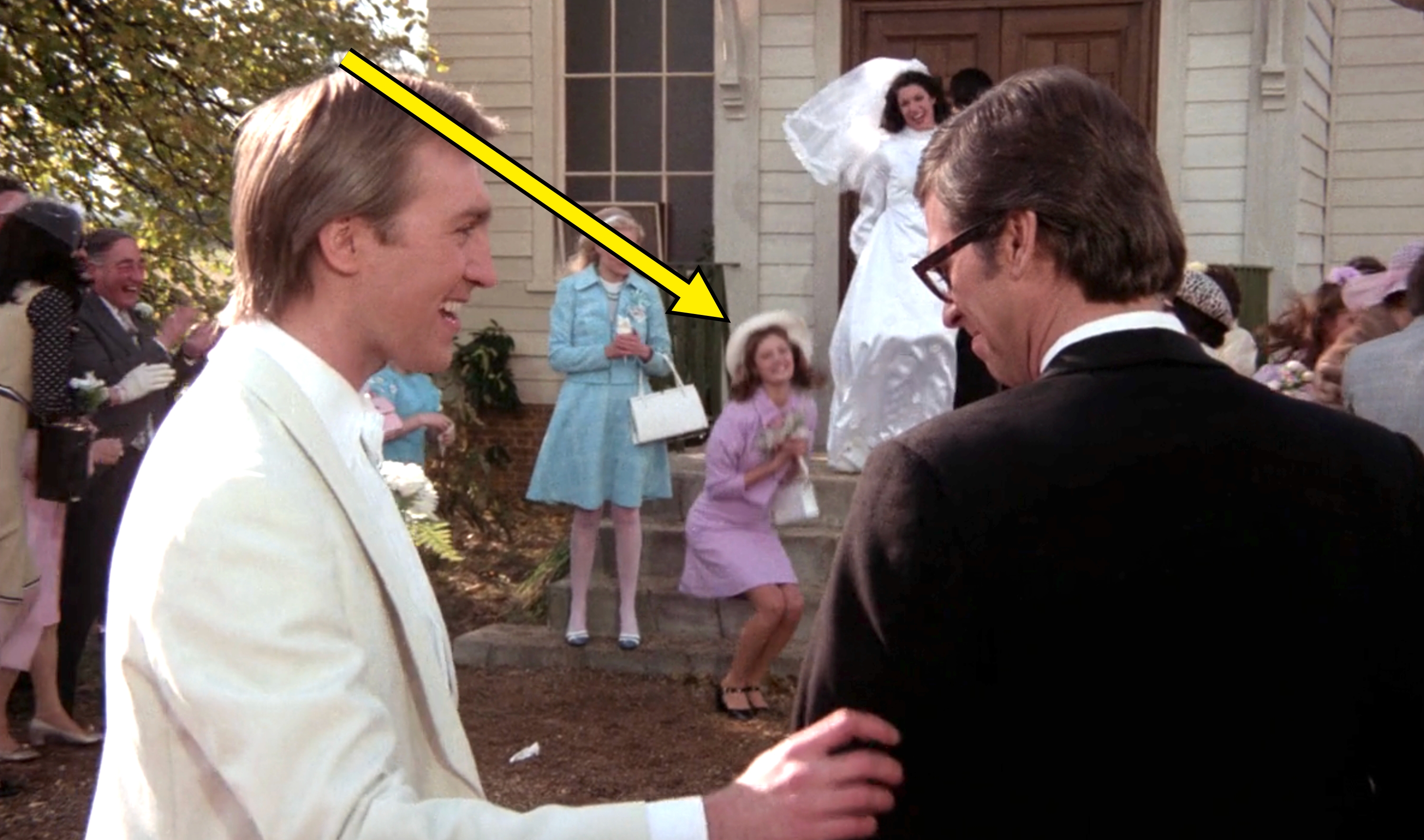 A happy groom in a white suit and glasses talks to a smiling man in a black suit at a wedding, with a bride and guests celebrating nearby