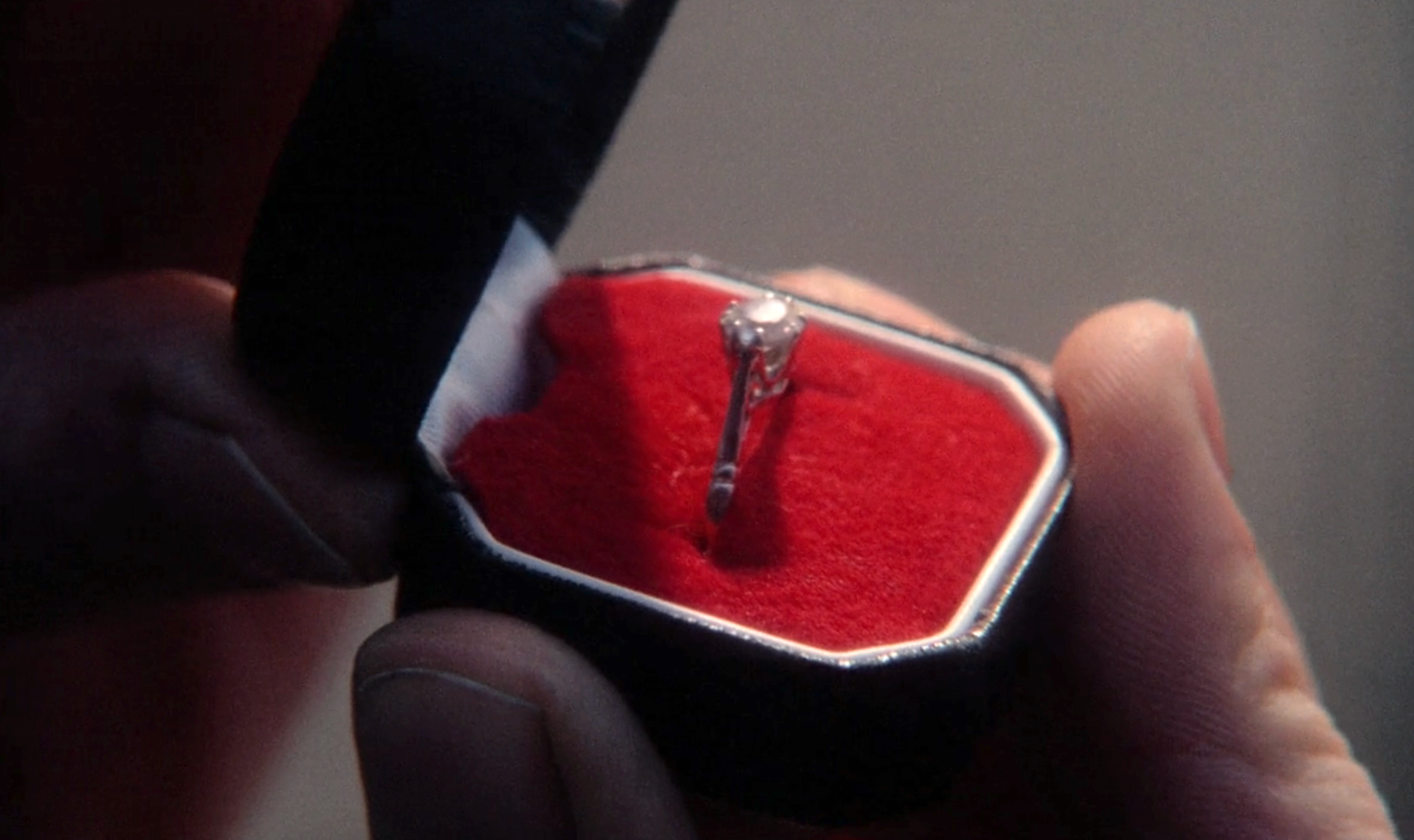 A person holds an open ring box with a diamond engagement ring set on a red cushion