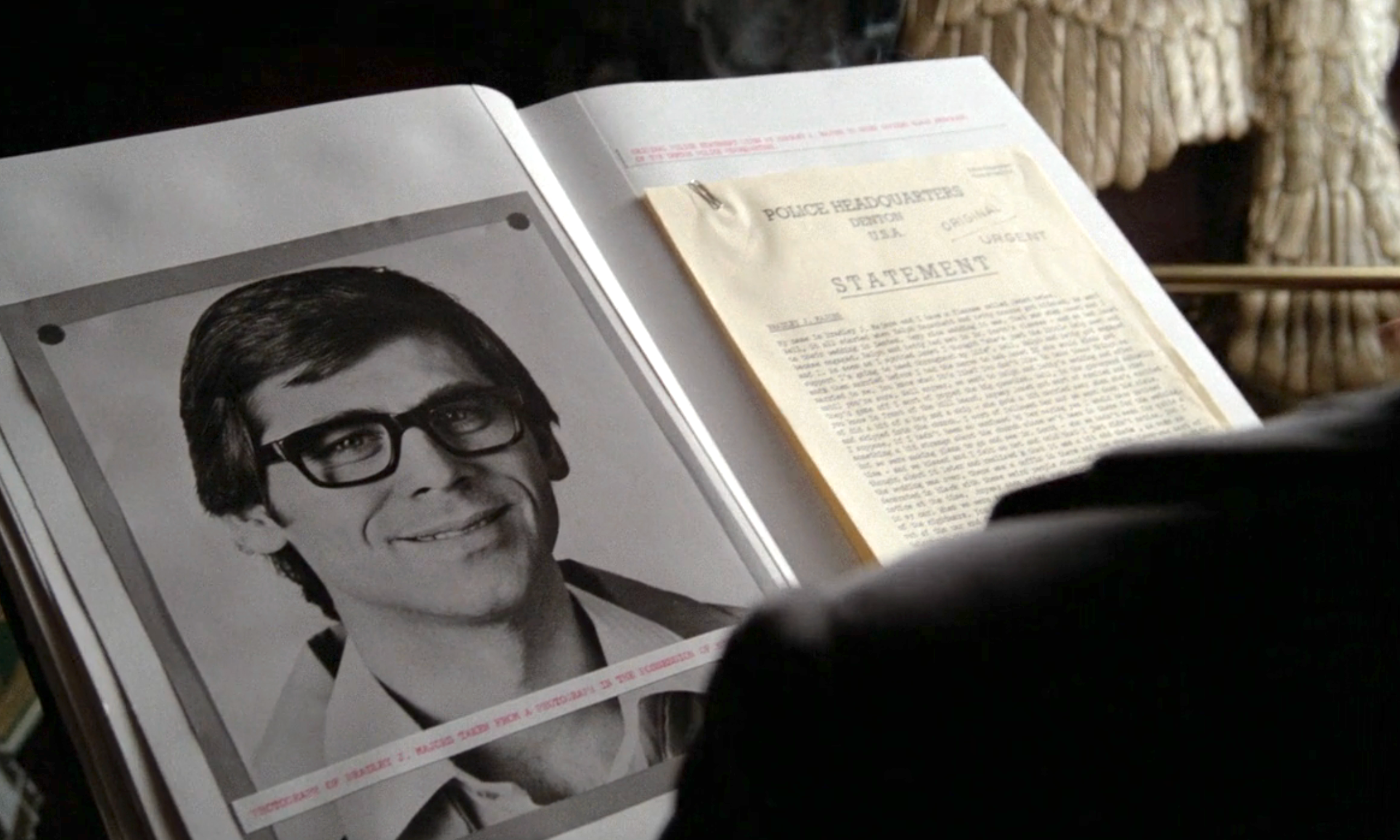 A black-and-white photo of a man with glasses is in a document next to a typed statement on a desk