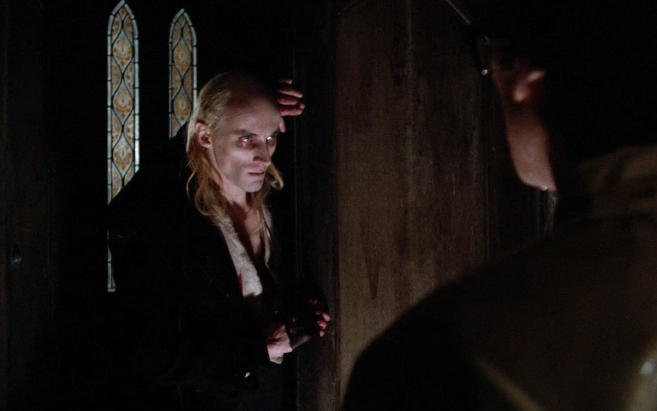 A gaunt figure with slicked-back hair stands in a dimly lit room, staring intensely at another person facing away from the camera