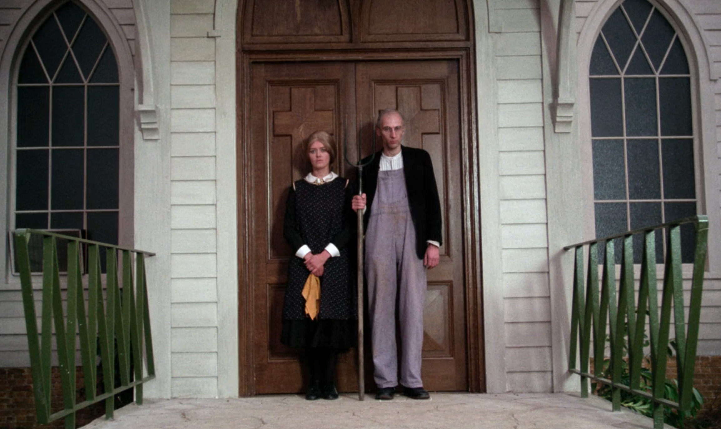 Two individuals stand in front of a wooden door, styled in early 20th-century attire, resembling an American Gothic aesthetic