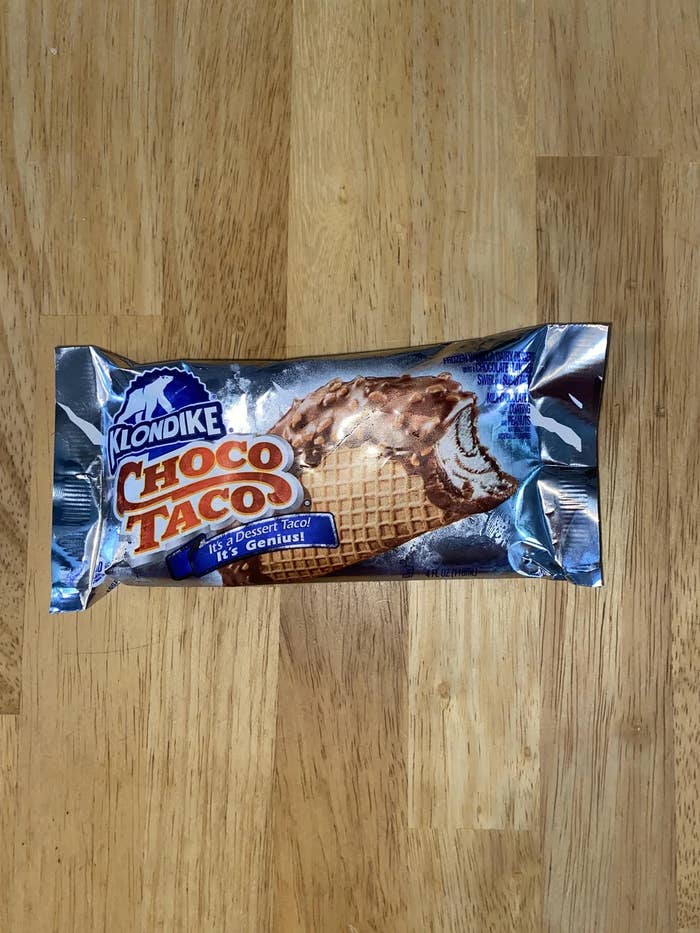 A wrapped Klondike Choco Taco ice cream treat on a wooden surface. The label reads "It's Dessert Taco! It's Genius!"