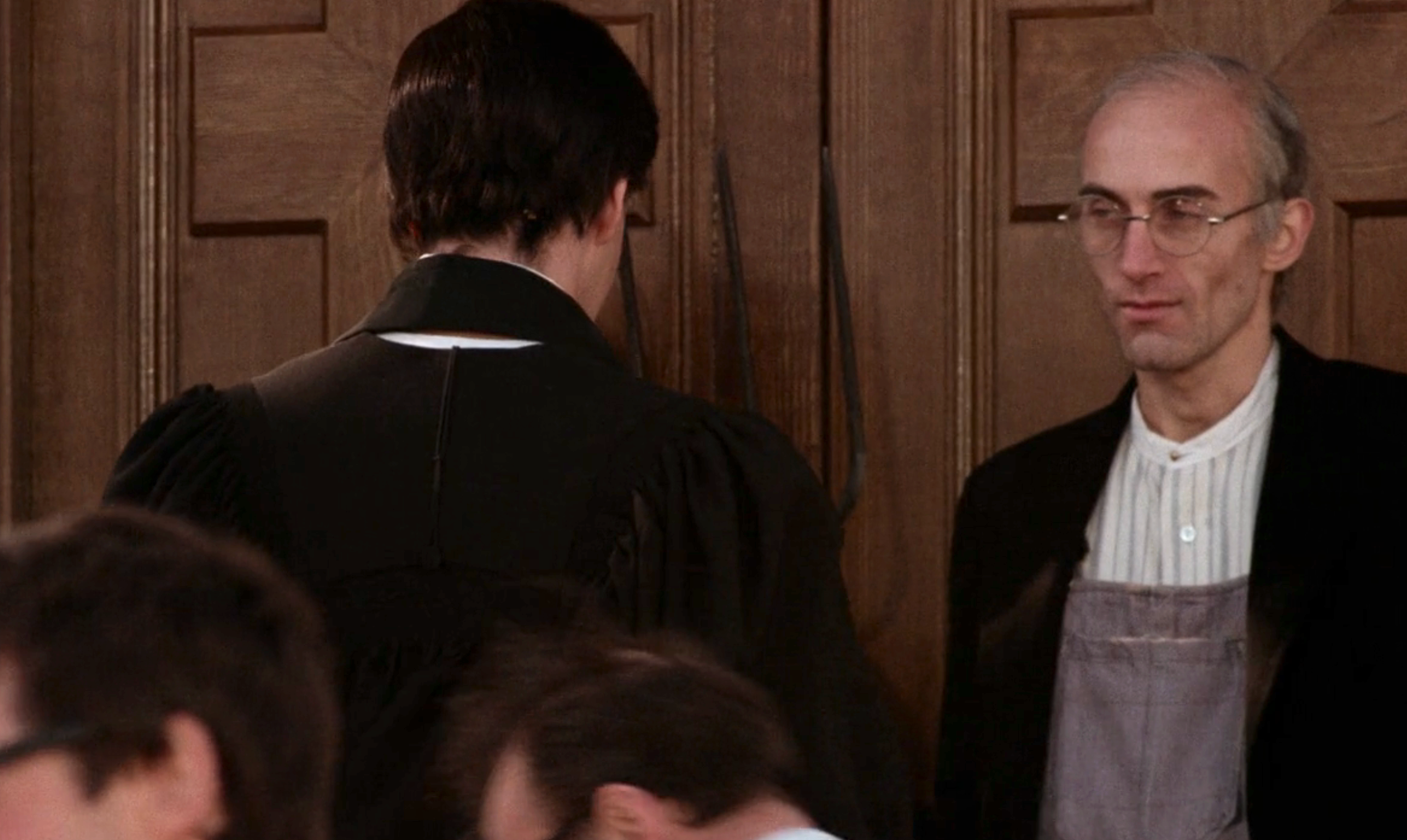 Two men wearing robes stand by a wooden door, facing away. One man looks on seriously