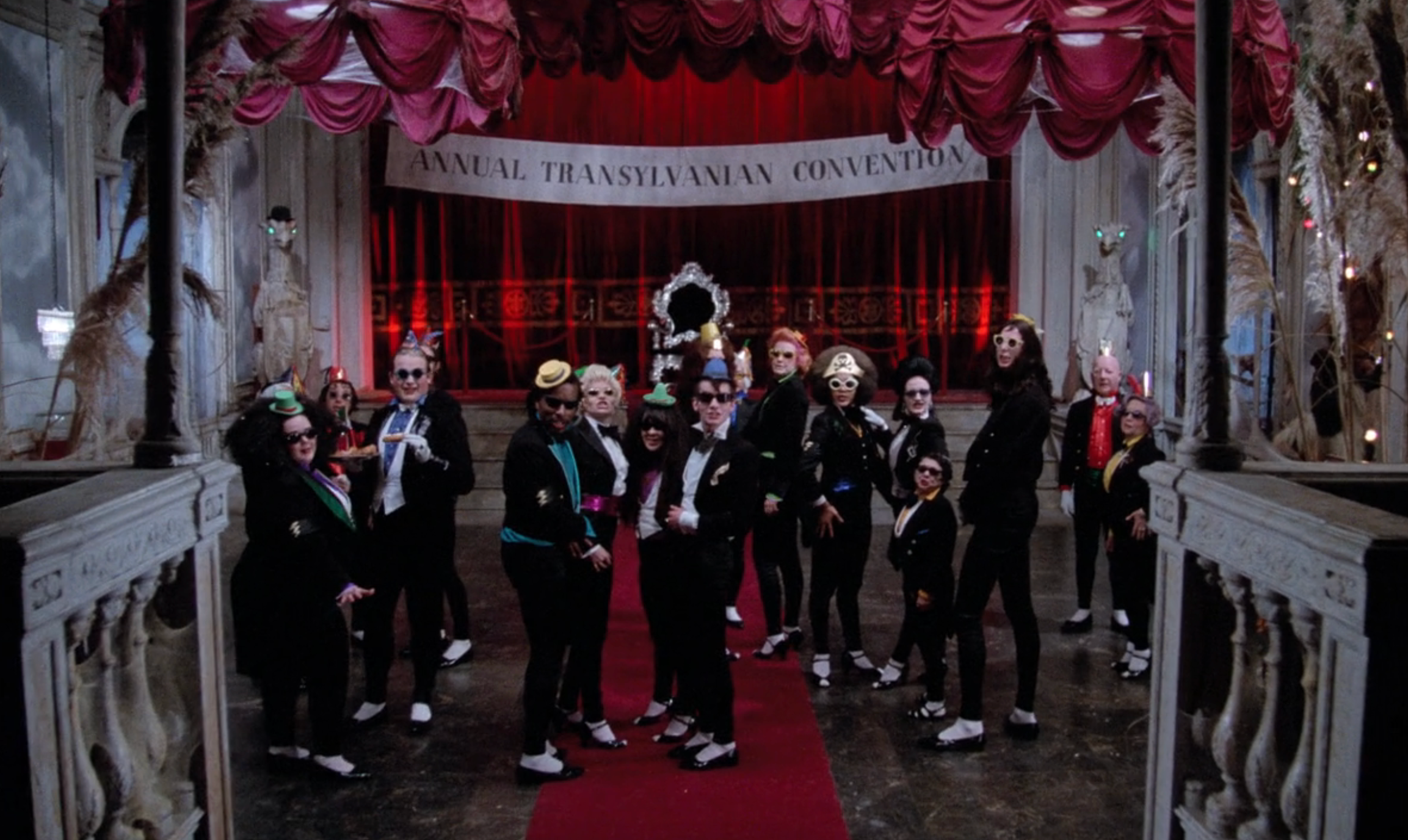 Group of people in formal and eccentric costumes at the "Annual Transylvanian Convention" with a red carpet and ornate backdrop