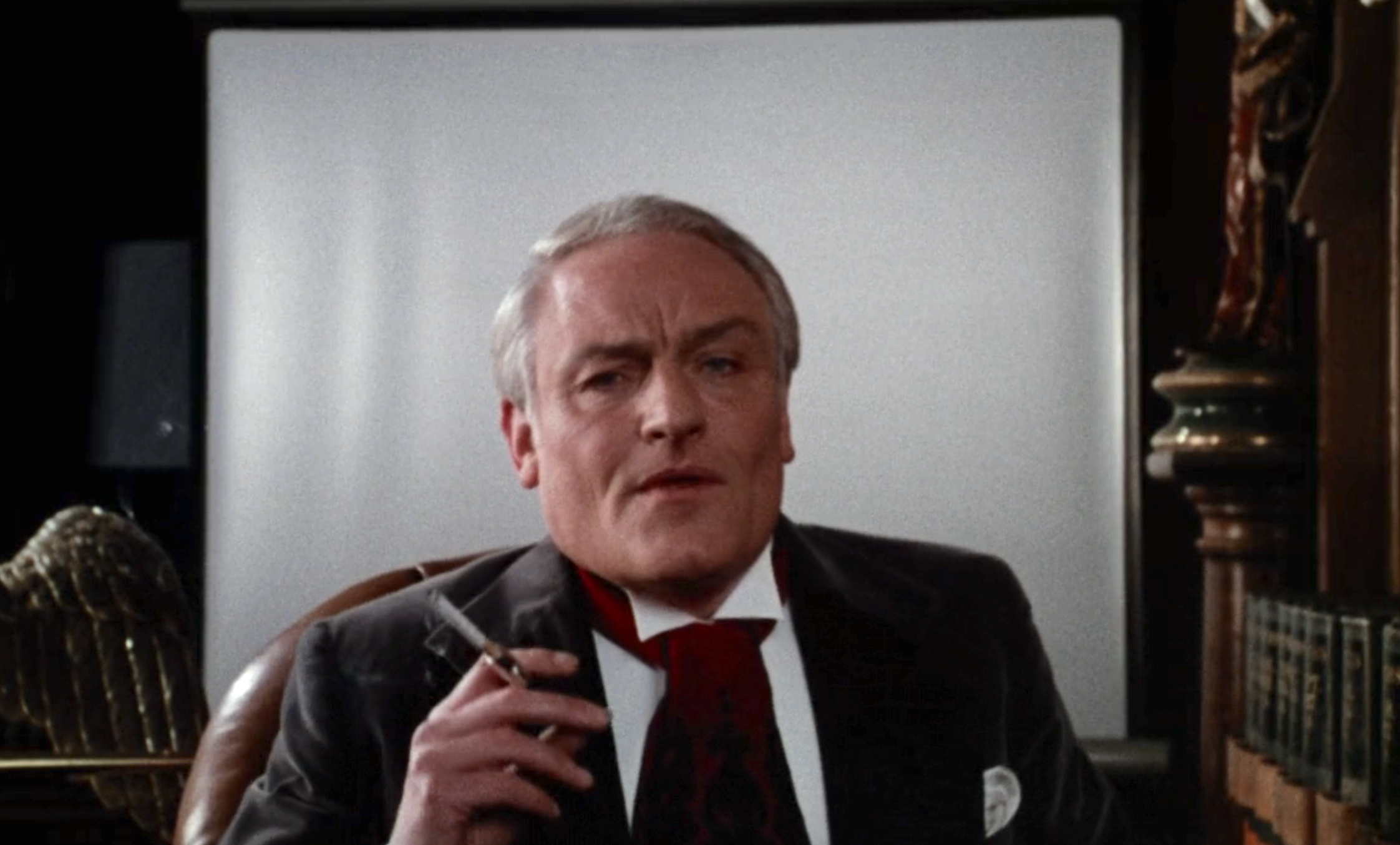 An older man in a suit sits in a chair, holding a cigarette. Behind him is a large screen or board