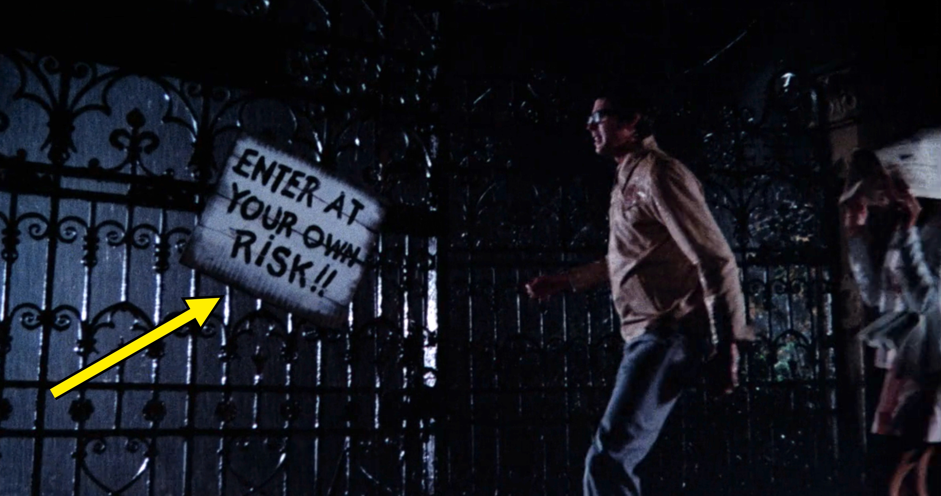 A man approaches an iron gate in the rain with a sign that reads "Enter At Your Own Risk!!"