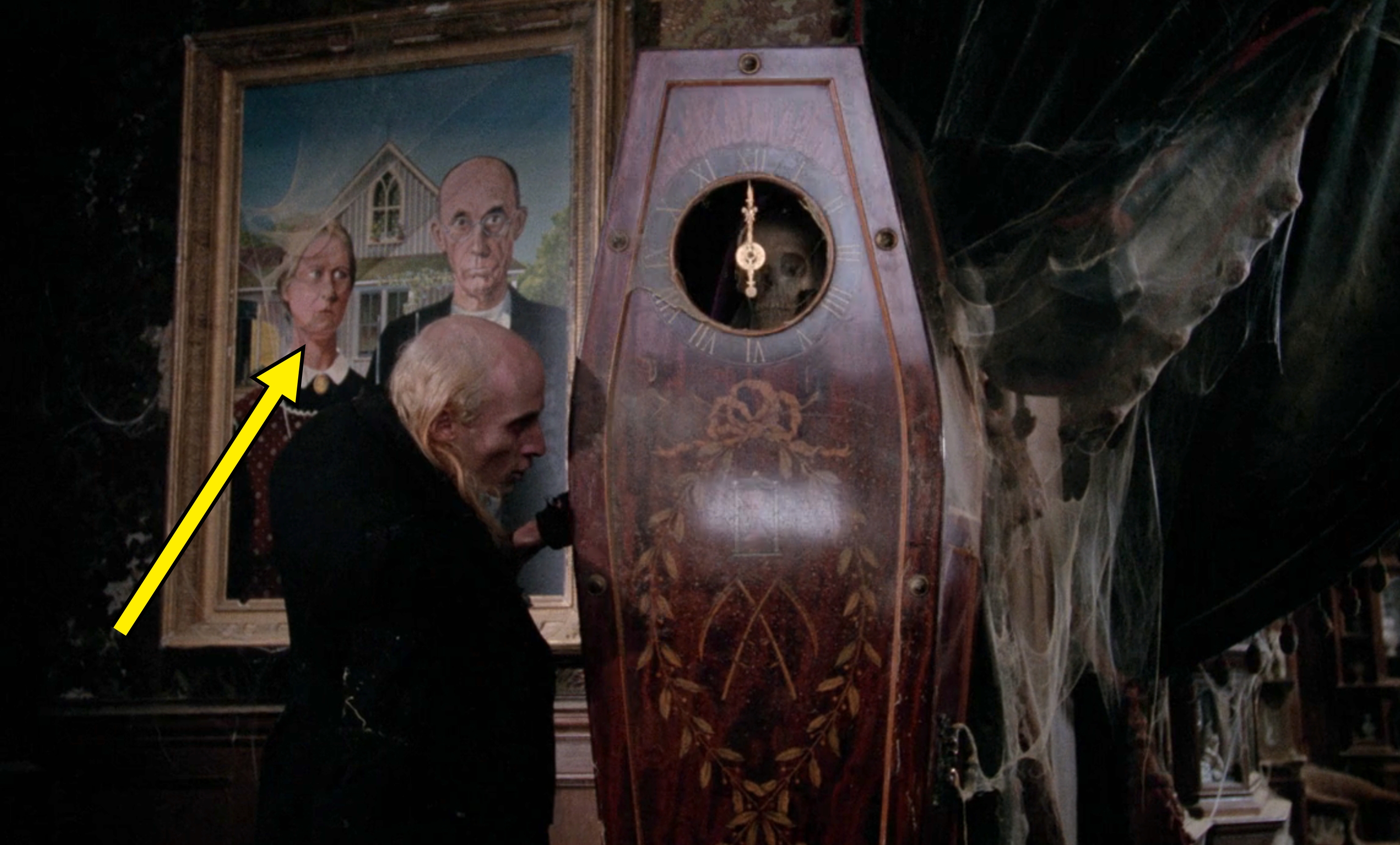 An elderly man examines a tall, ornate coffin-like clock in a dimly lit room with cobwebs; a painting of a stern couple is on the wall behind him