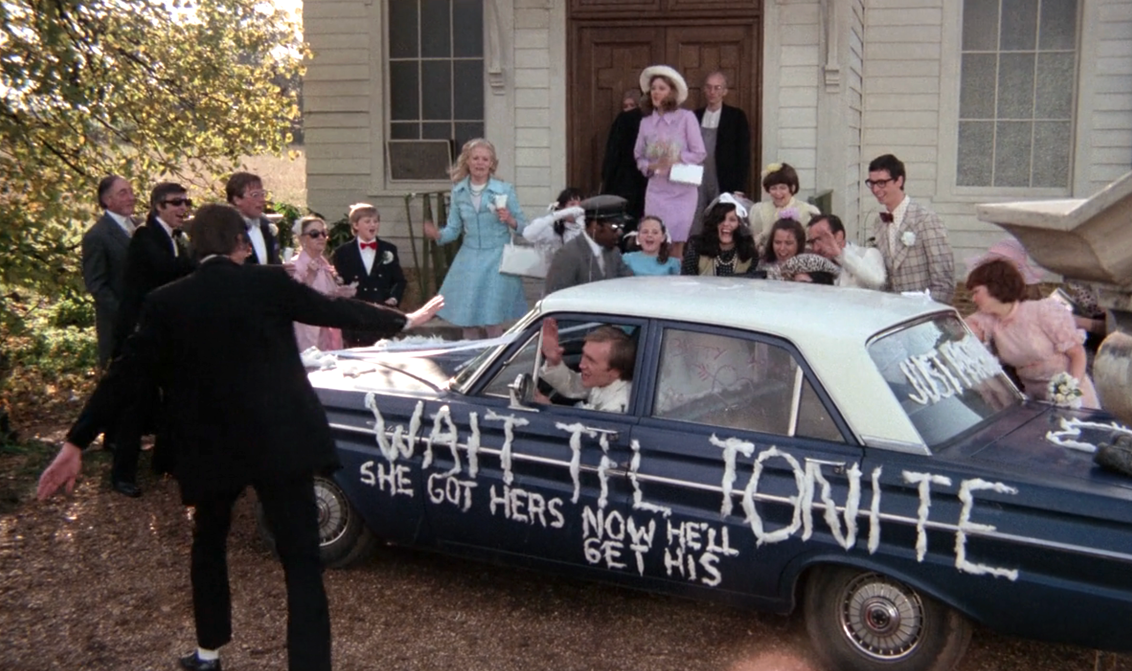 People around a car painted with "Wait Til Tonite She Got Hers Now He'll Get His" during a wedding scene in front of a church