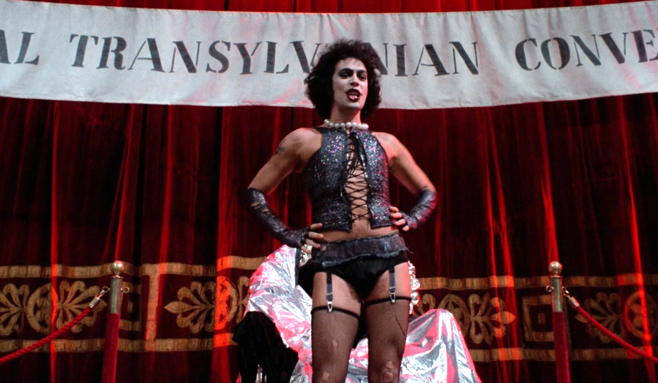 Person in theatrical costume with corset and fishnet stockings stands confidently on stage in front of a "Transylvanian Convention" banner