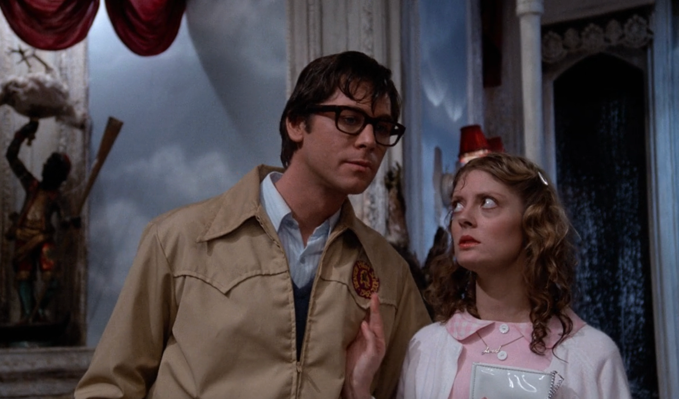 A man in glasses wearing a jacket and a woman with a curly hairstyle in a cardigan stand inside a vintage-style room, both with curious expressions