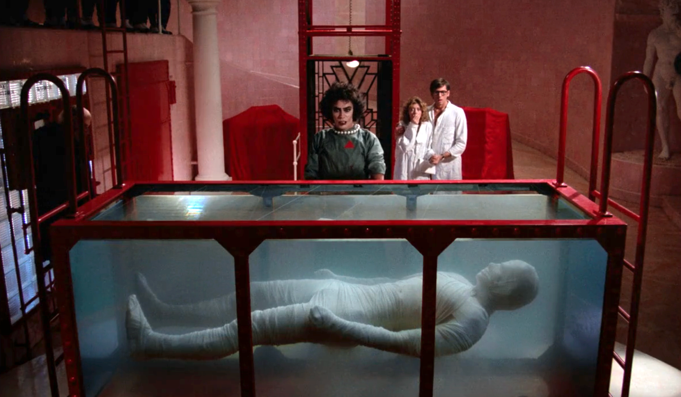 A scientist in a green outfit stands beside a transparent tank with a humanoid figure inside. Two people in white lab coats observe