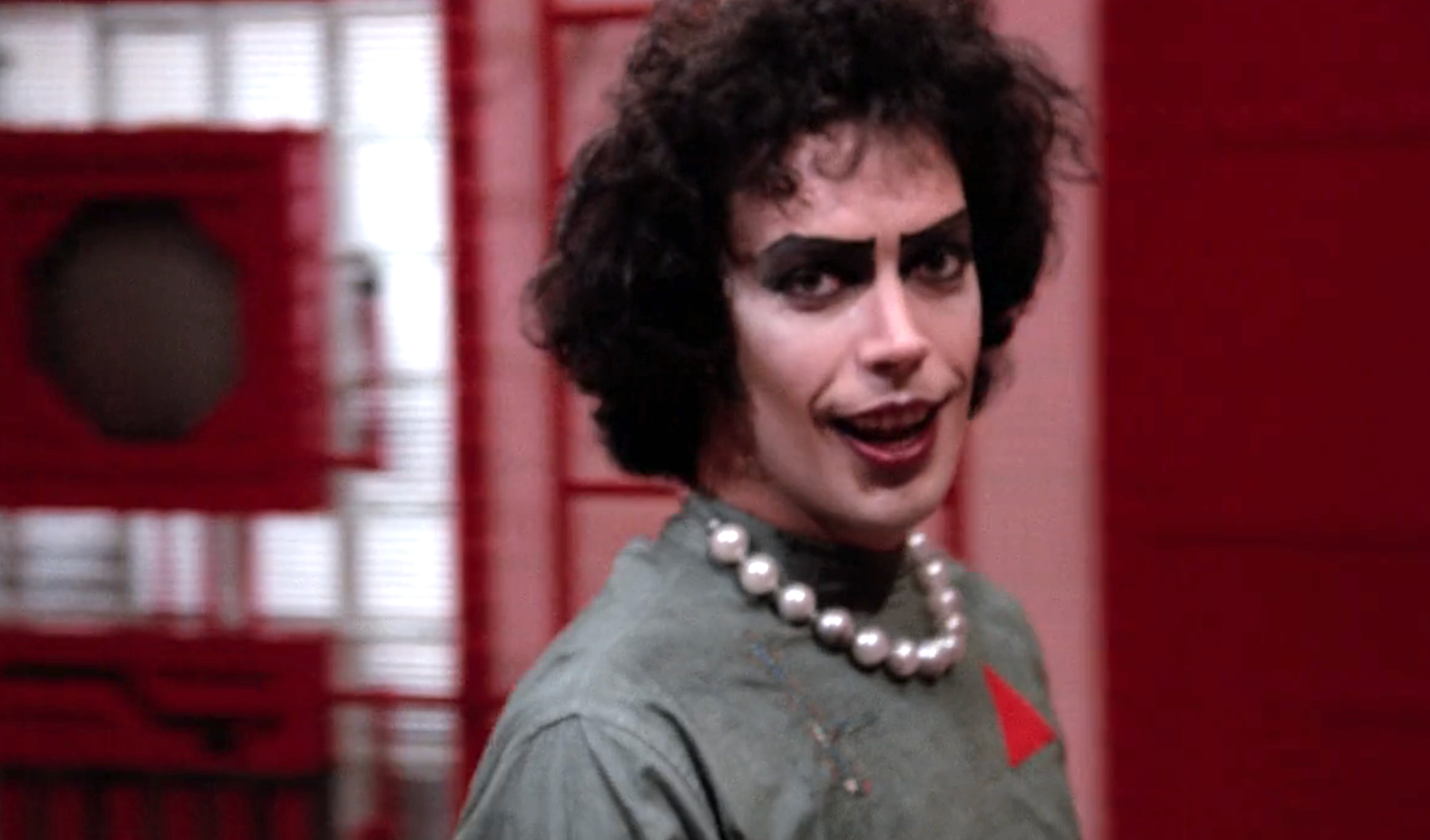 Person in dramatic makeup and pearls, wearing a costume with a theatrical flair, looking directly at the camera with a confident expression