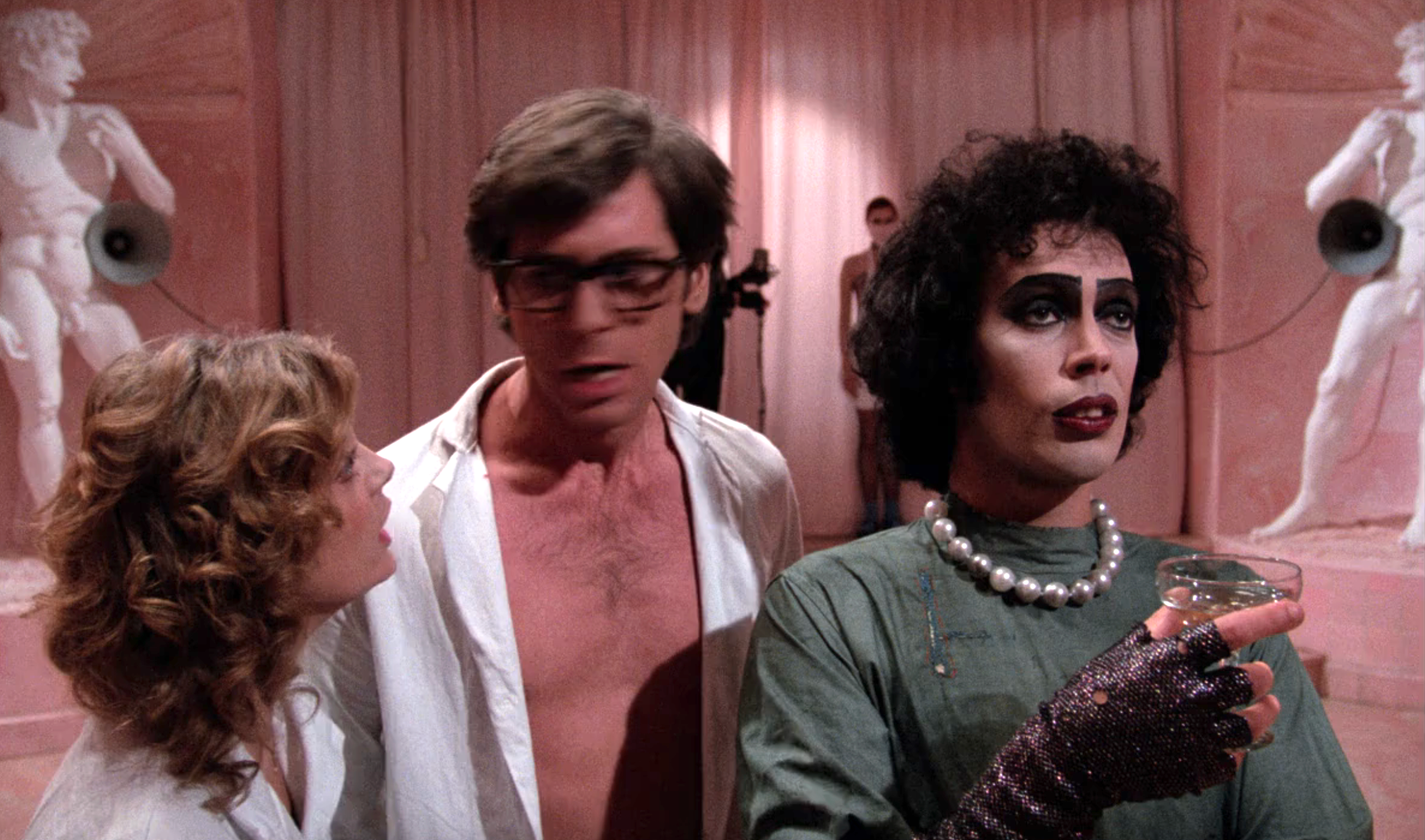Three people in a theatrical setting, one in dramatic makeup and pearls, another shirtless with glasses, and a third looking surprised