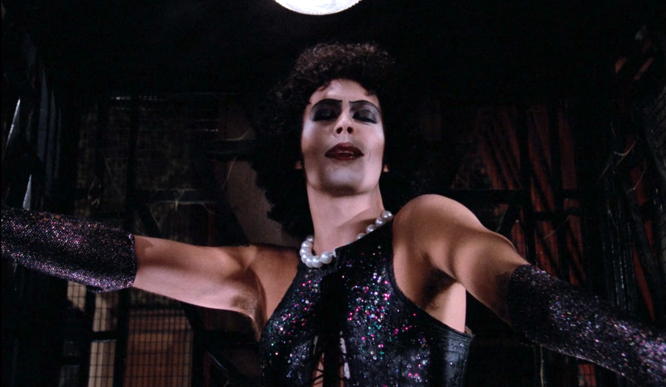 Person in dramatic makeup and glittery outfit with arms outstretched, wearing a pearl necklace, in a theatrical setting