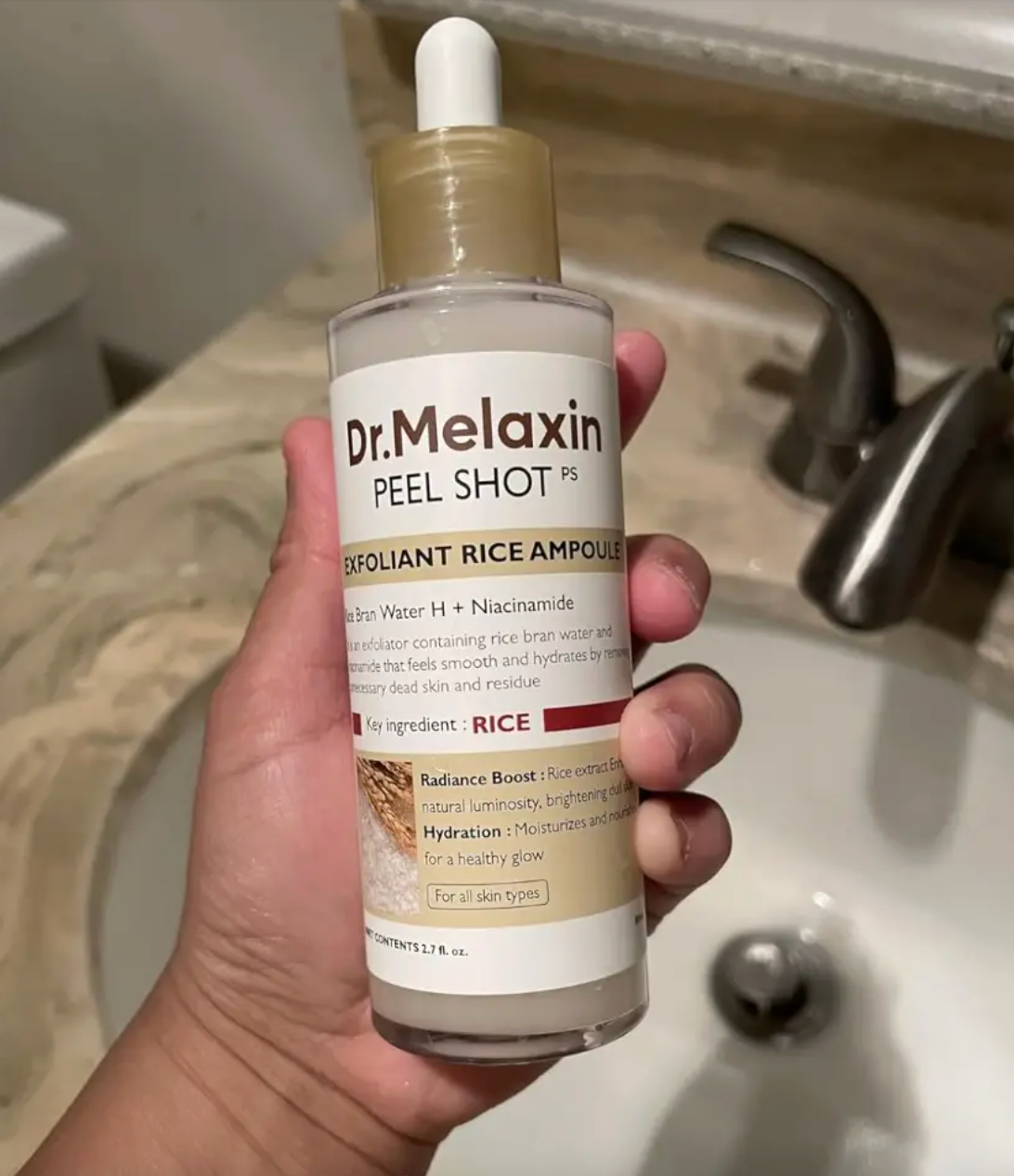 A manus  holding a vessel  of Dr. Melaxin Exfoliant Rice Ampoule by a sink, highlighting its atom   and niacinamide ingredients for tegument  hydration and radiance