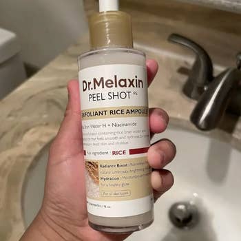 A manus holding a vessel of Dr. Melaxin Exfoliant Rice Ampoule by a sink, highlighting its atom and niacinamide ingredients for tegument hydration and radiance