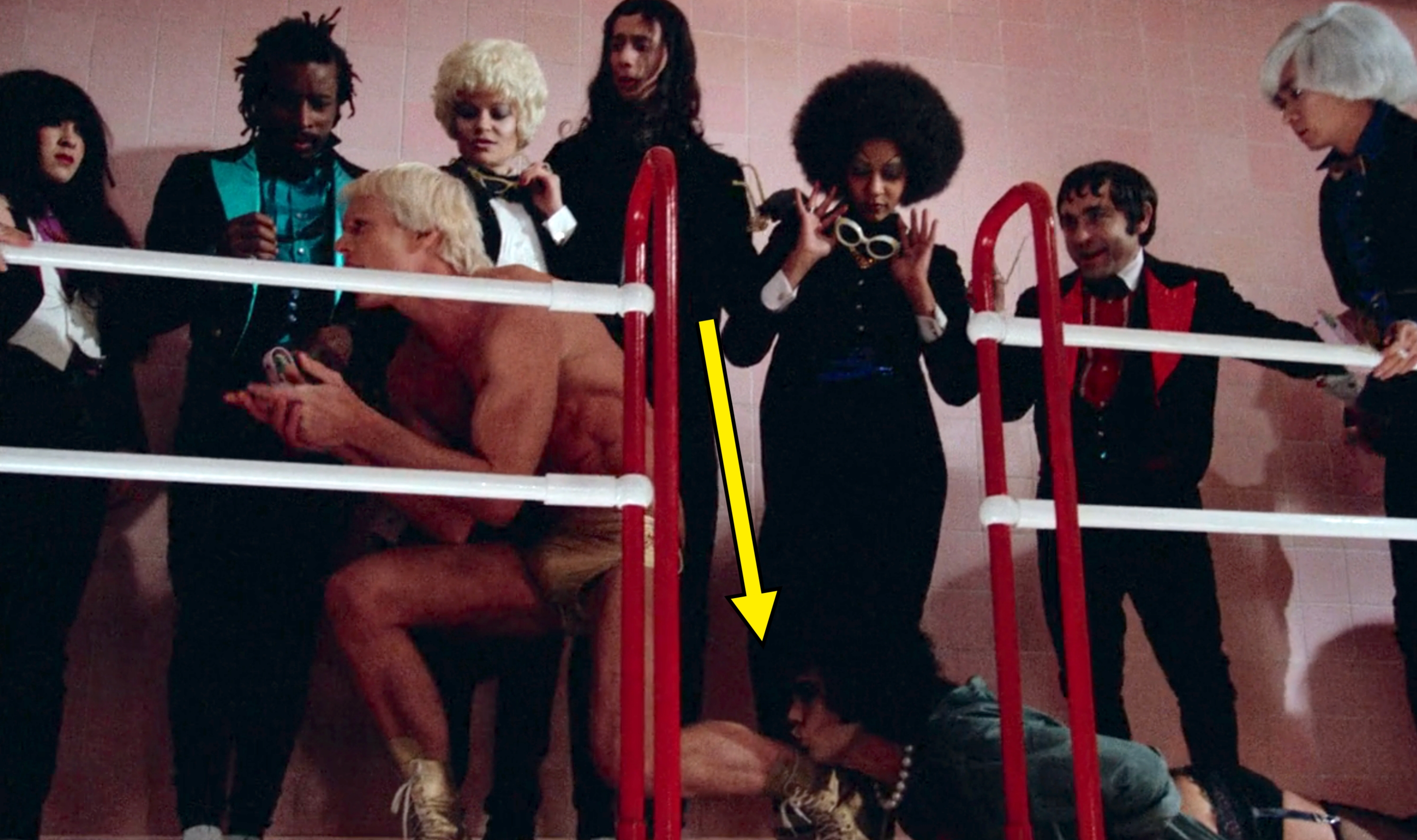 People in costume and wigs gathered around a man wearing only shorts inside a boxing ring. The scene has a theatrical and surreal style