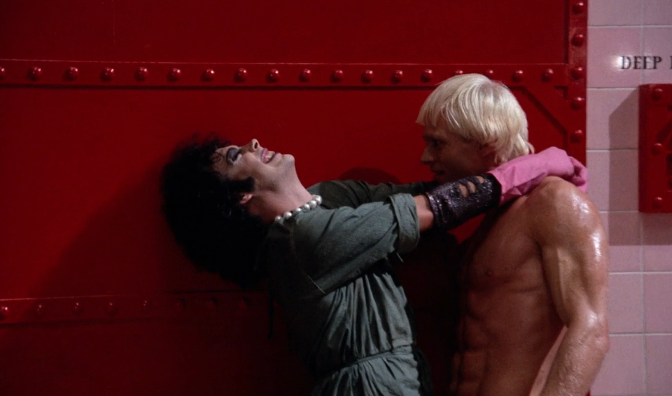 Two people in a theatrical pose, one with dark hair and dramatic makeup wearing pearls, the other shirtless with short blonde hair, set against a red wall