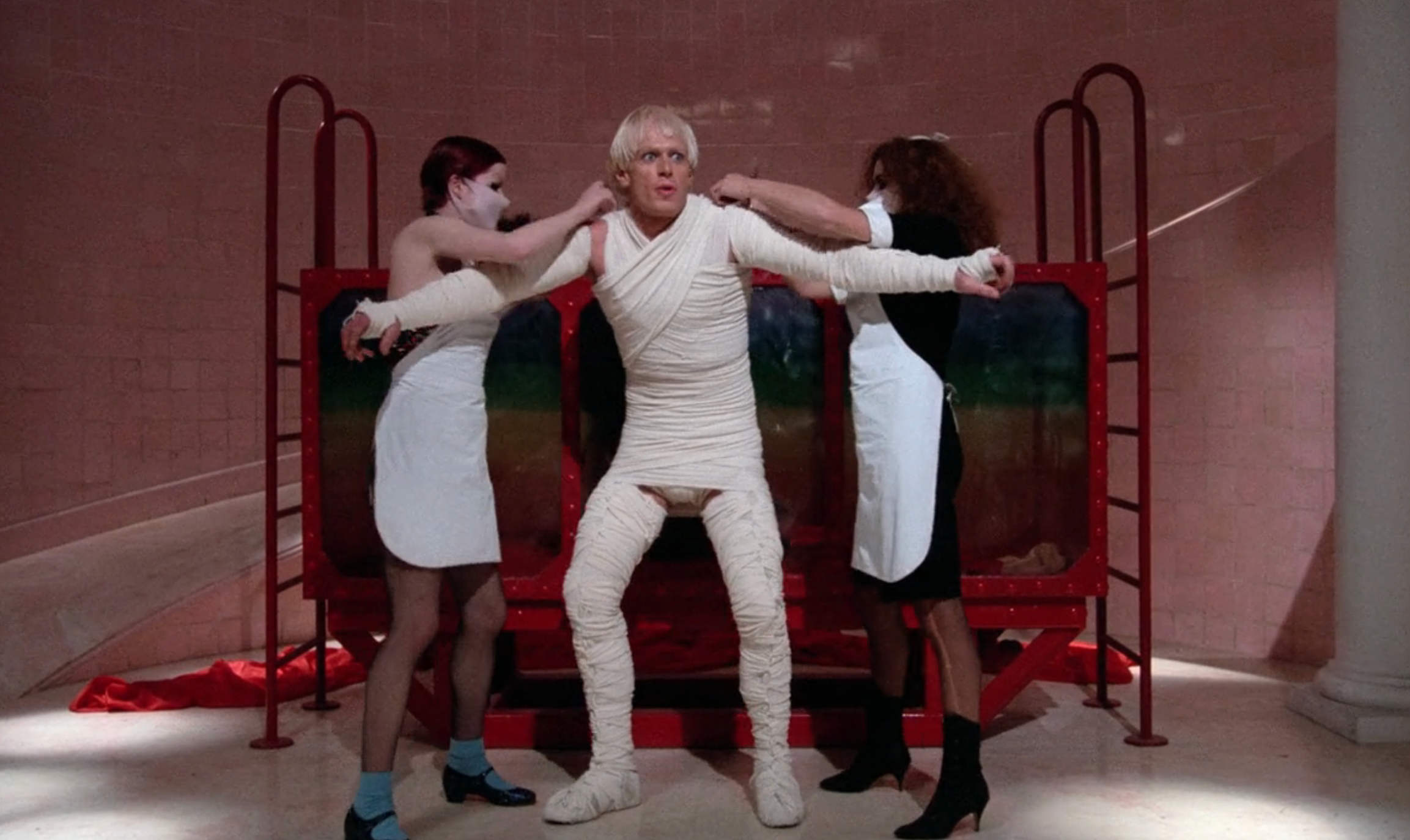 Person wrapped in bandages, assisted by two masked individuals in a staged, theatrical setting