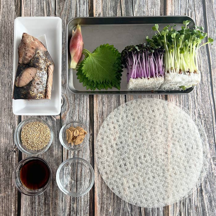 料理の材料セット。焼き魚、薬味、大葉、新鮮な野菜、ライスペーパーなどが食器に盛られている。