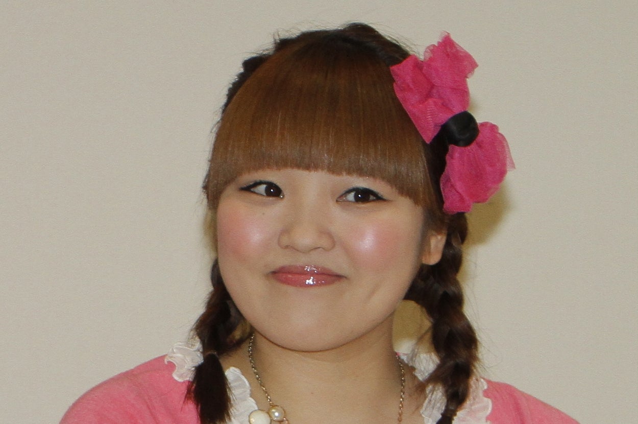 著名人の女性が花を手に笑顔。髪は編んであり、大きな花飾りがついています。カジュアルで華やかな服装です。