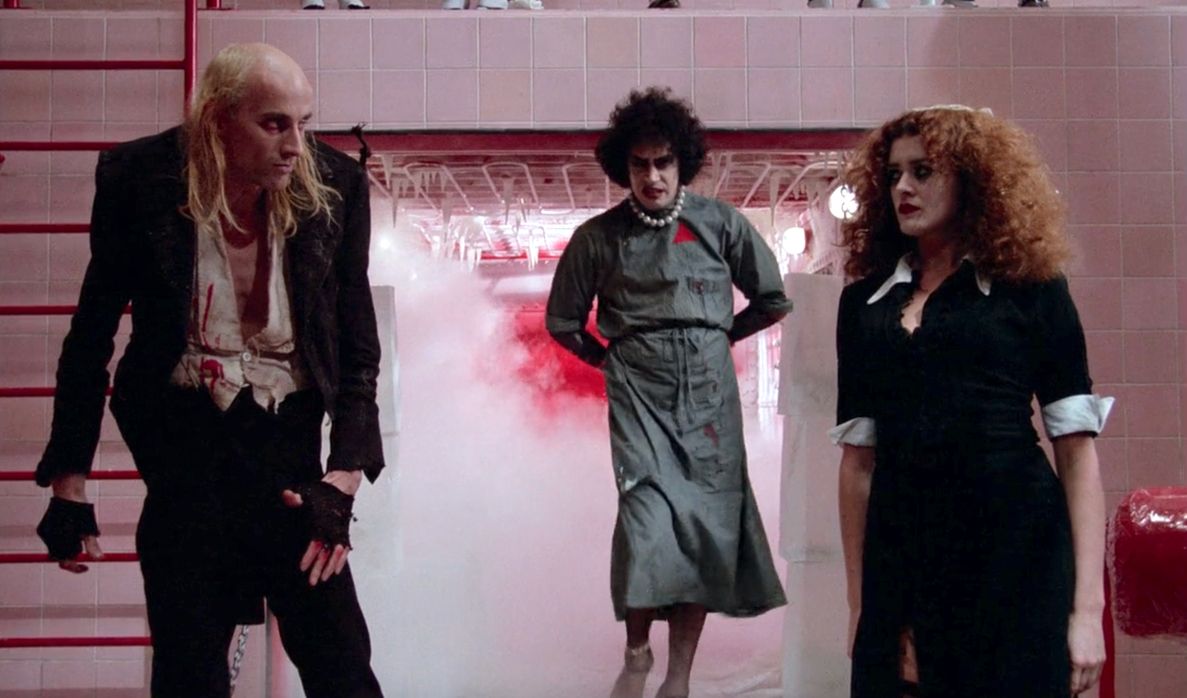 Three actors in theatrical costumes and makeup stand in a misty, dramatic setting, conveying a sense of tension and intrigue