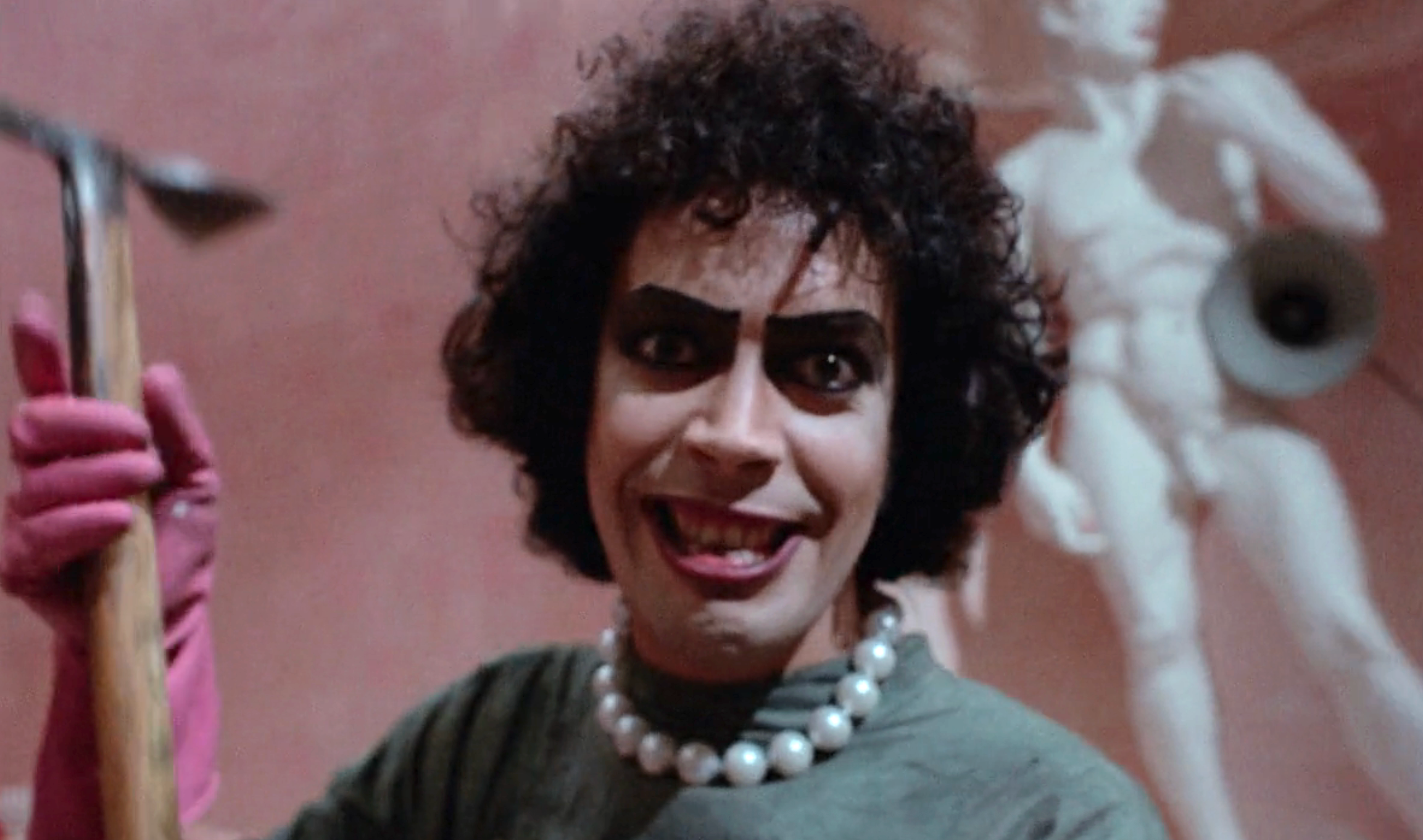 Person with curly hair, pearls, dark eyebrows, wearing gloves, holding a tool, with a statue in the background, smiling energetically