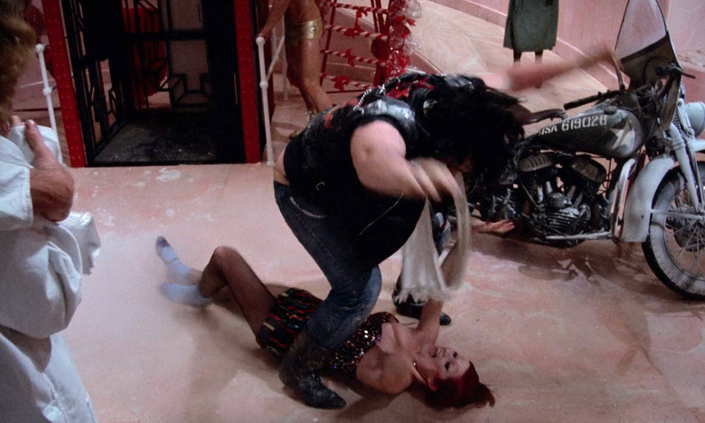 Person wrestling on floor in a theatrical scene, with a motorcycle backdrop. Another person in costume looks on