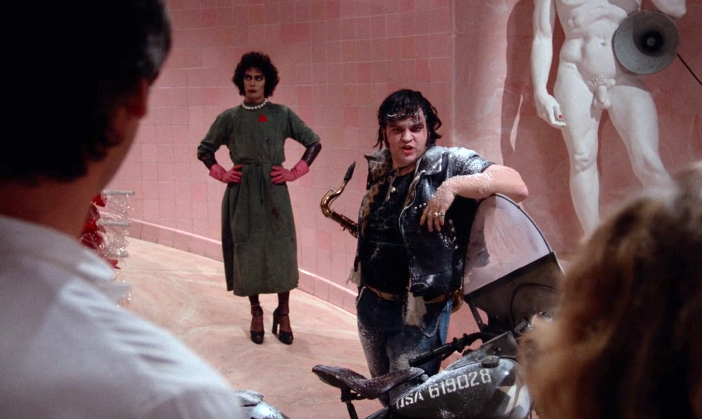 A person in a dress watches another in a leather outfit pose dramatically beside a motorcycle in a pink-tiled room