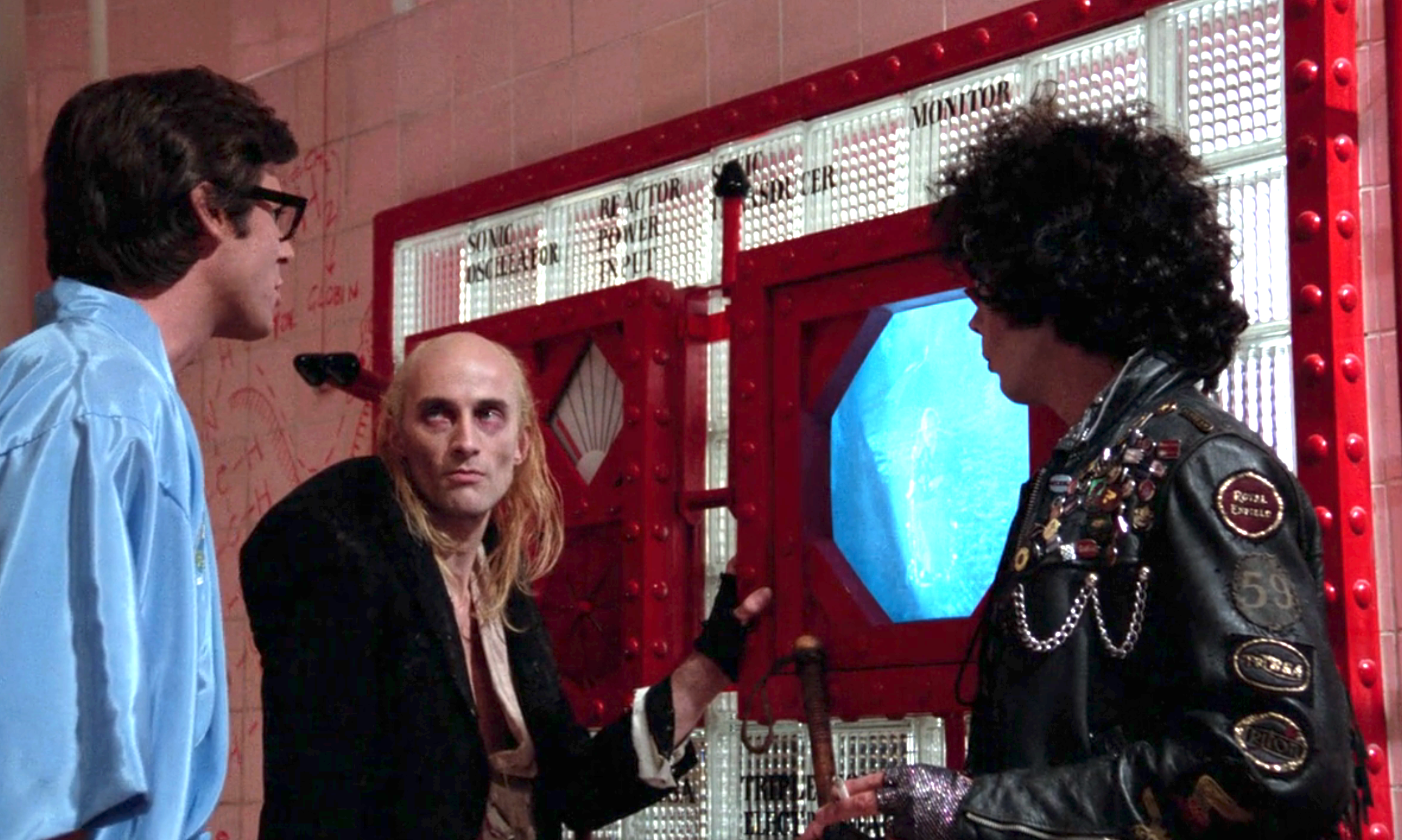Three people in a futuristic room with one opening a door to a glowing panel. The person on the right wears a leather jacket with numerous patches