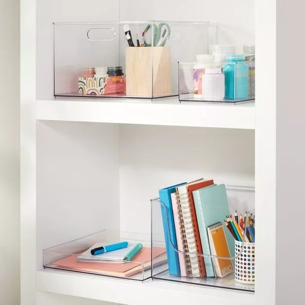 Tempat penyimpanan bening di rak putih menampung perlengkapan seni, buku, dan alat tulis, ideal untuk kantor rumah atau ruang belajar yang terorganisir