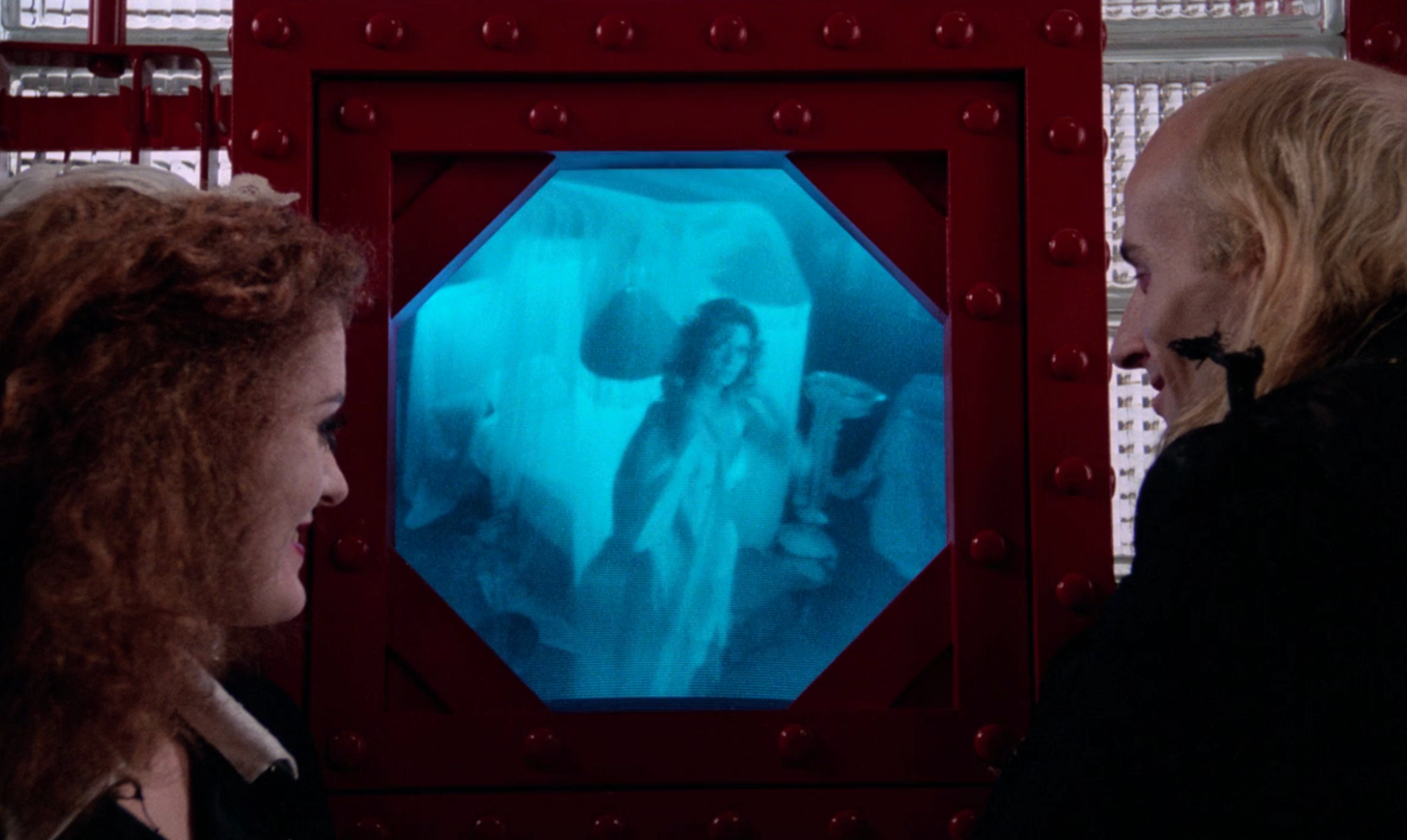 Two people look at a holographic image displayed in a futuristic setting, the hologram shows a woman in a flowing gown