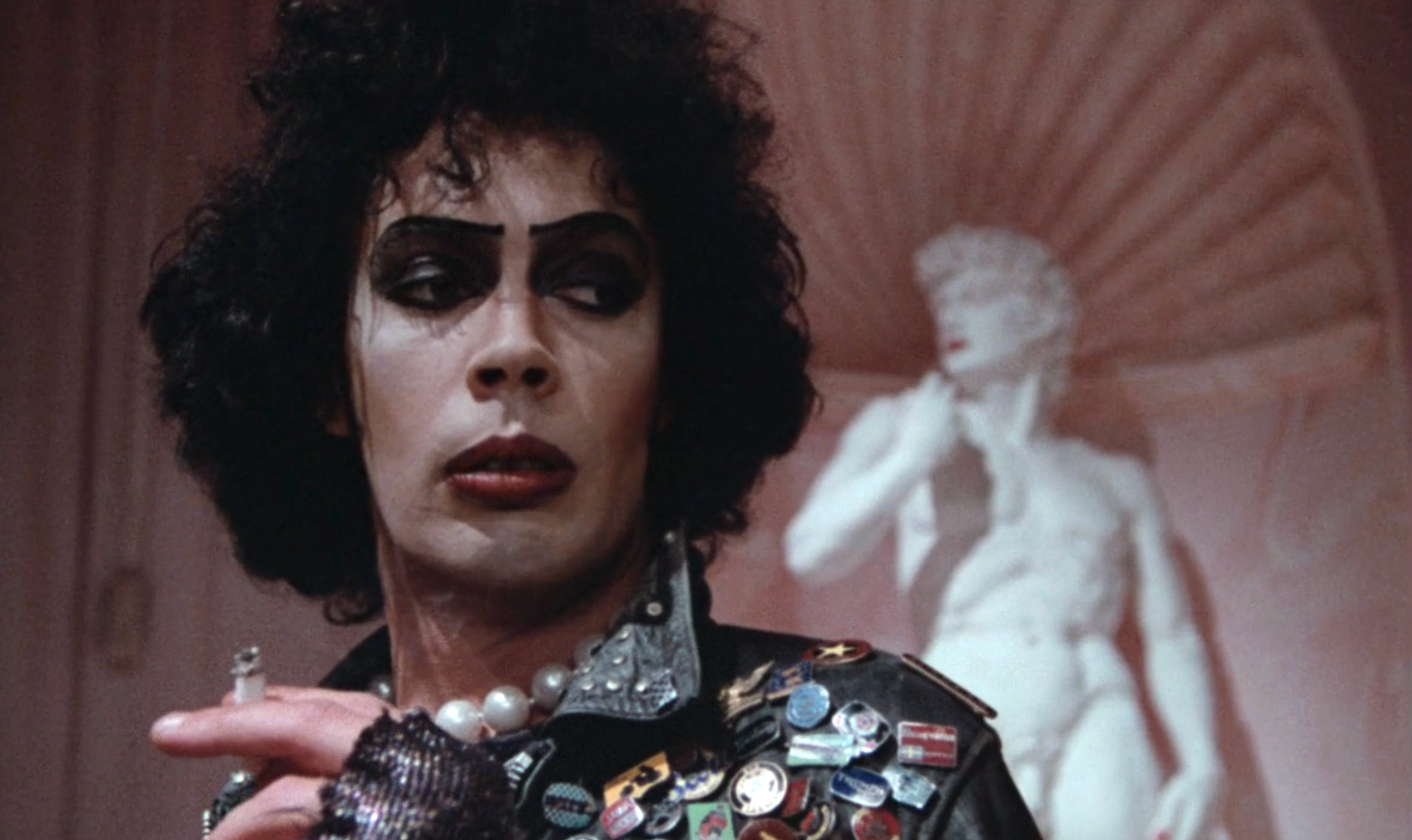 Person in dramatic makeup and leather jacket with pins, standing indoors. Marble statue in the background