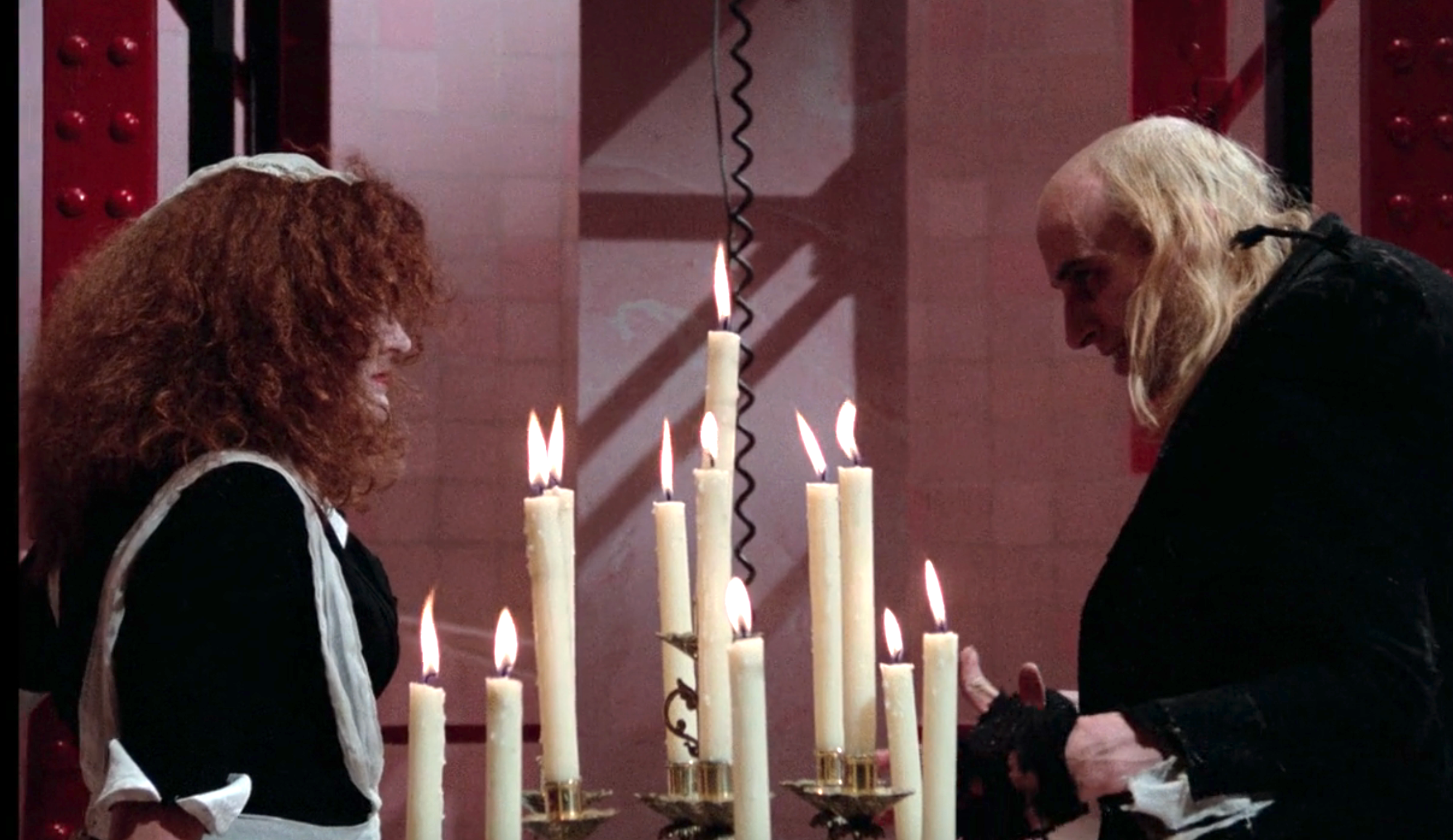 Two people dressed in costume, one as a maid and the other with a bald cap and long hair, stand facing each other across a table with lit candles