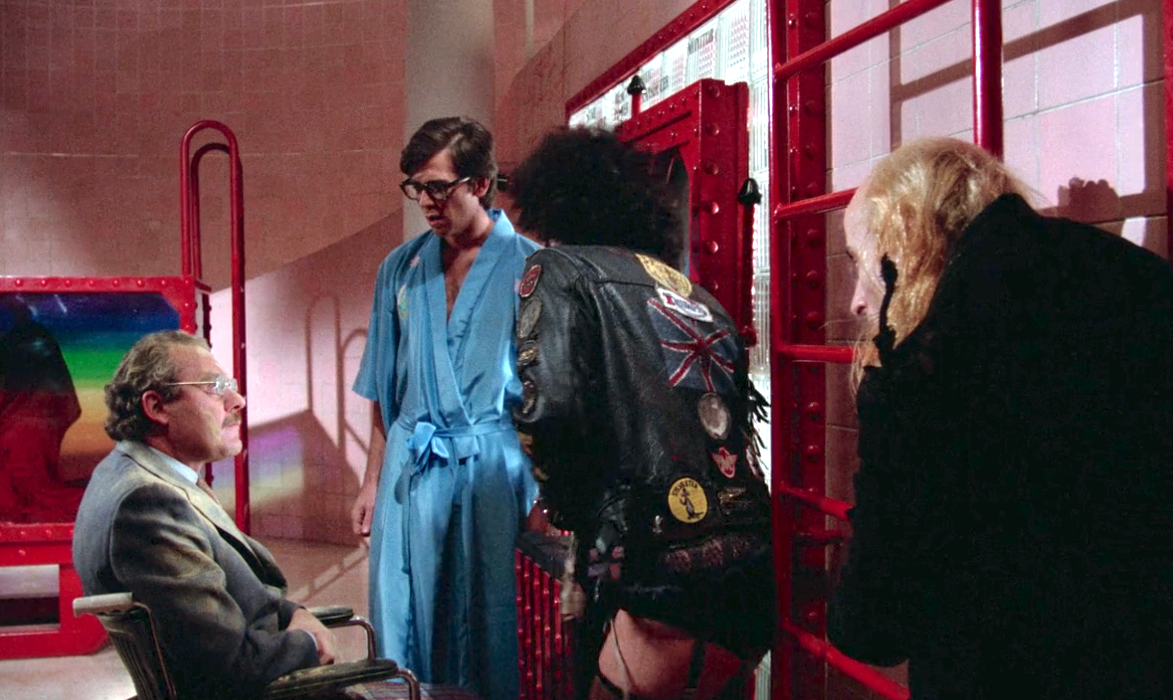 A group of people in a room; one person in a blue robe talks to a seated man, while others in eclectic outfits engage nearby