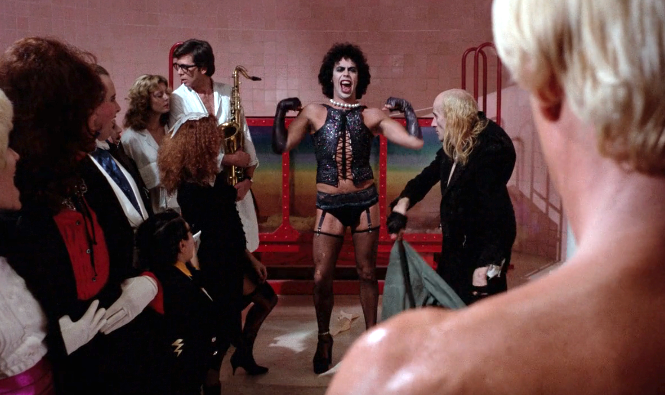 A performer in a corset and stockings is surrounded by people, some in costumes. One holds a saxophone. Another person in leather is in the foreground